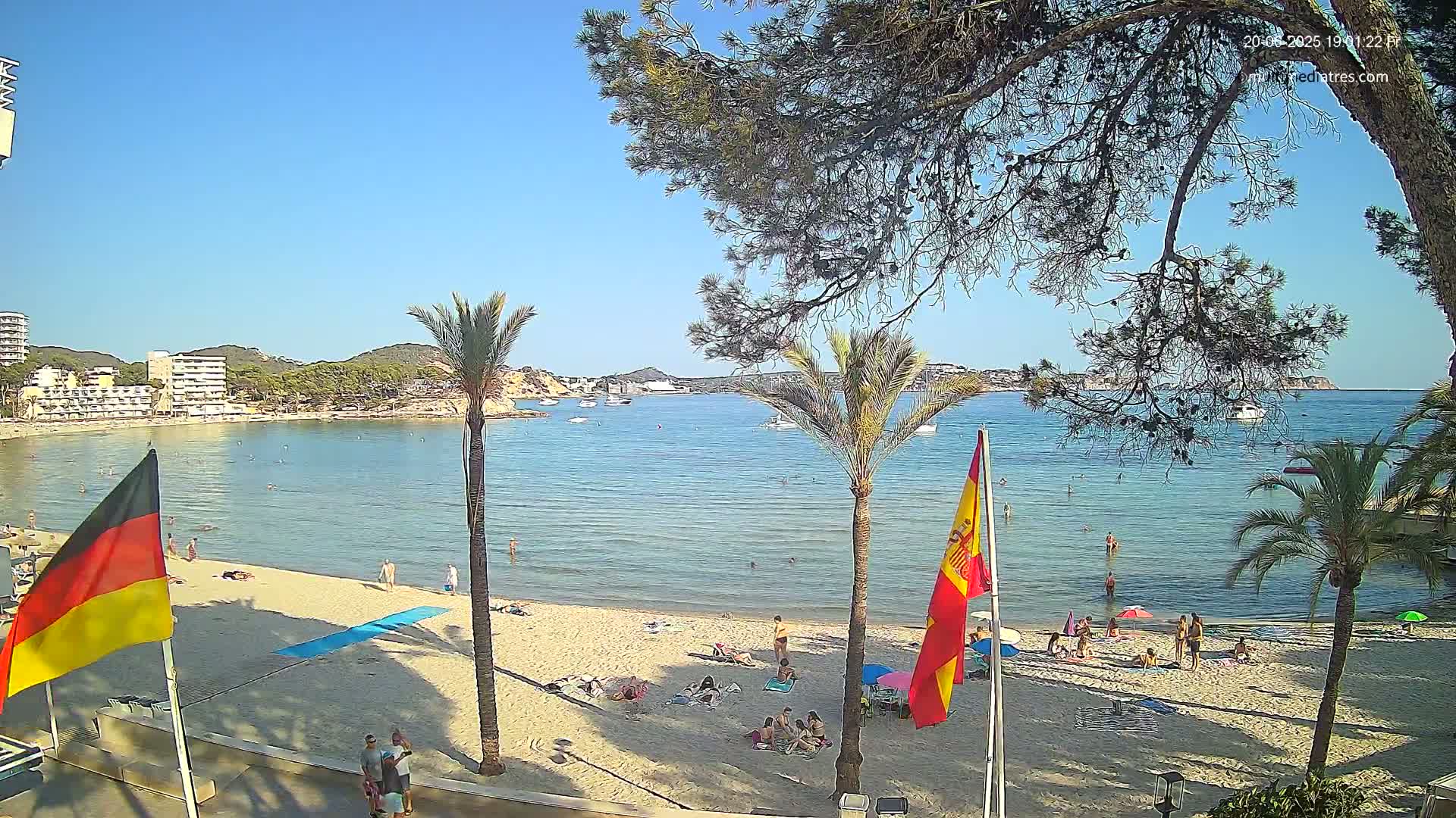 A sandy beach with calm, clear water, palm trees, and people sunbathing and swimming under a sunny sky.