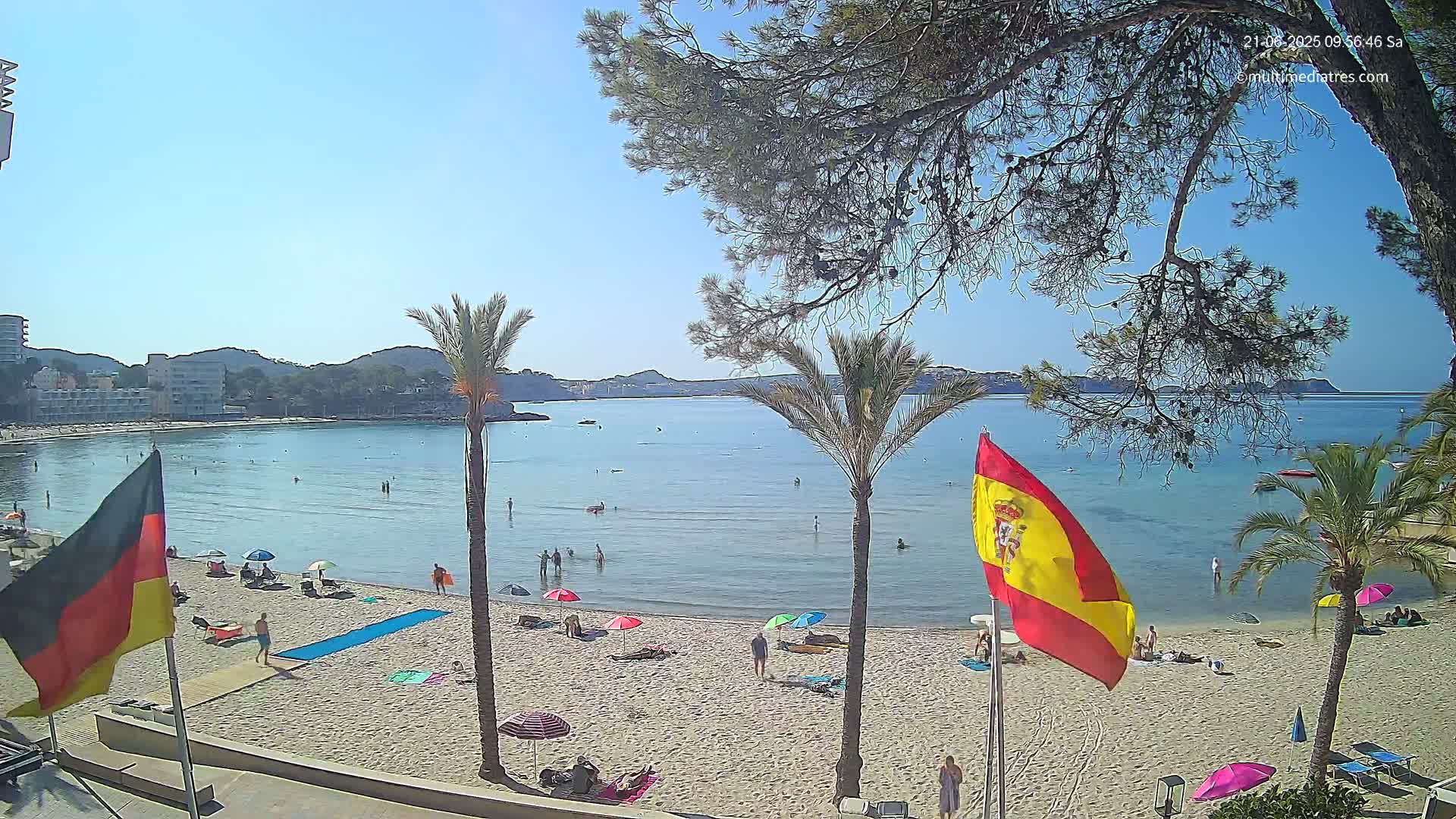 A sandy beach with calm, clear water and several people swimming and sunbathing, under a sunny sky, is bordered by palm trees and buildings.