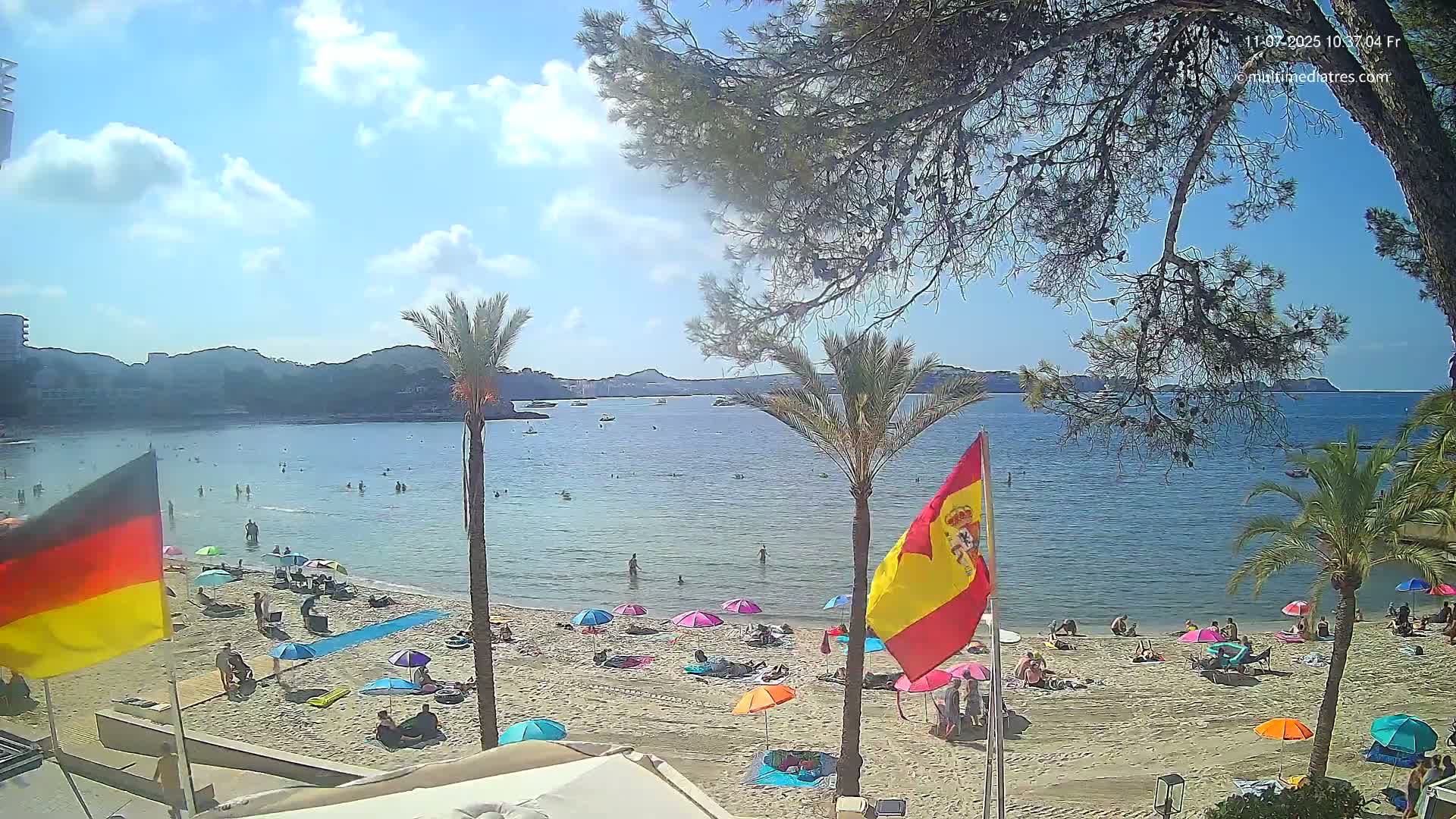 A sunny beach scene shows a calm bay with people swimming and sunbathing under colorful umbrellas, palm trees, and flags on a sandy shore.