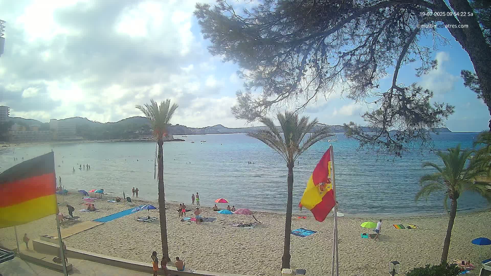 A partly cloudy day at a beach with calm water, showing several people swimming and relaxing on the sand under umbrellas, with palm trees and flags visible.