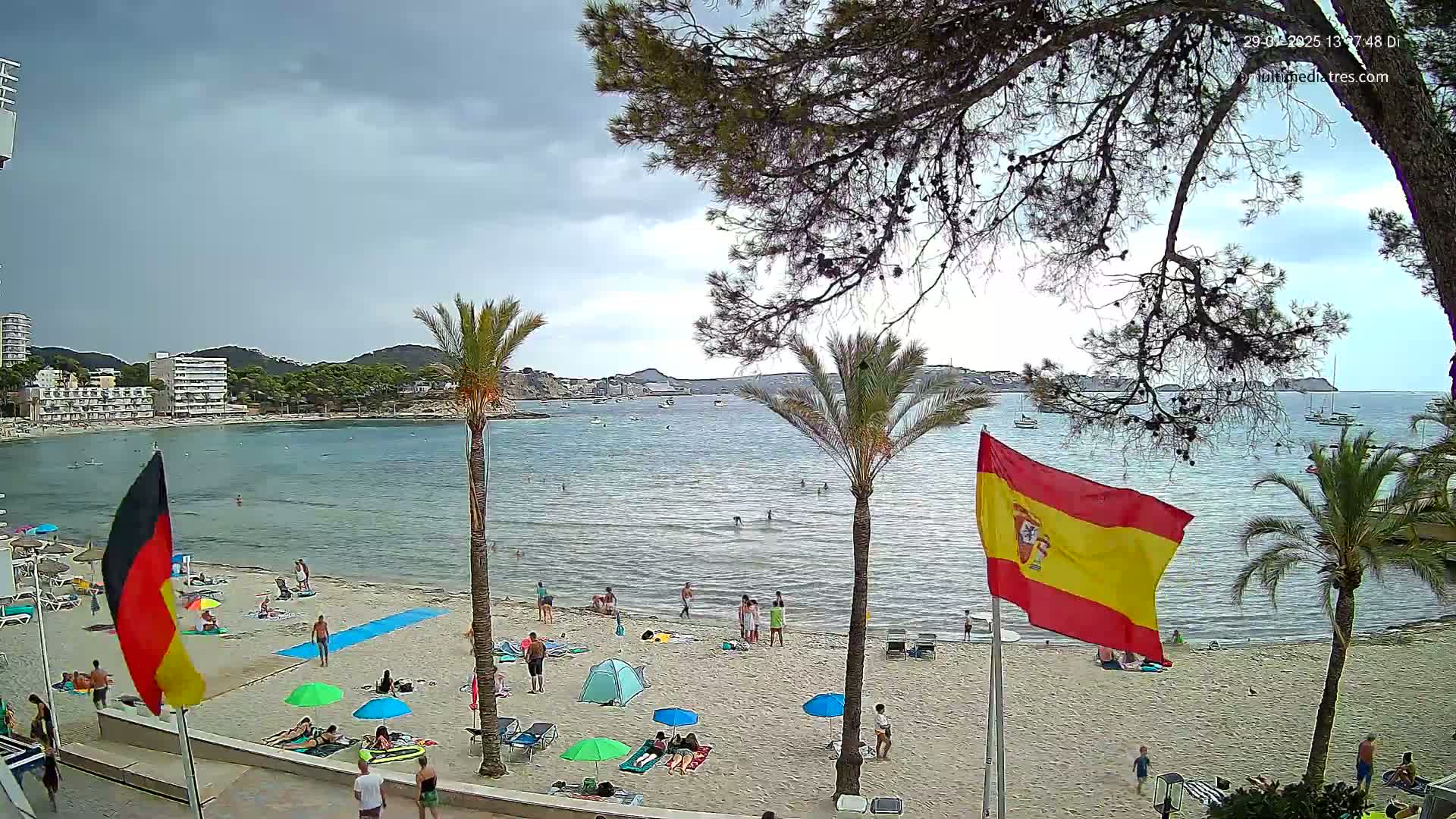 A partly cloudy day at a beach with calm water, showing several people swimming and relaxing on the sand under umbrellas, with palm trees and flags visible.