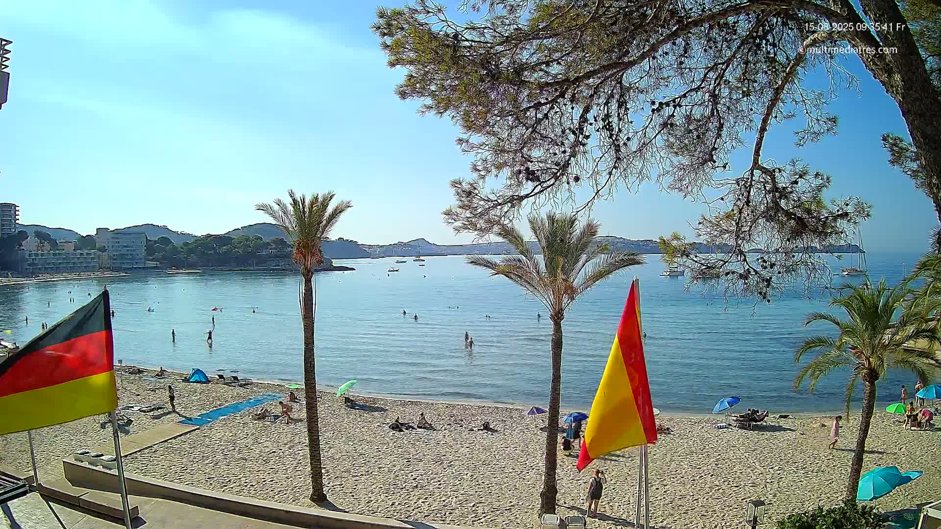 A sunny beach scene shows a sandy shore with people swimming and sunbathing, palm trees, and boats on a calm, clear blue bay, under a mostly clear sky.