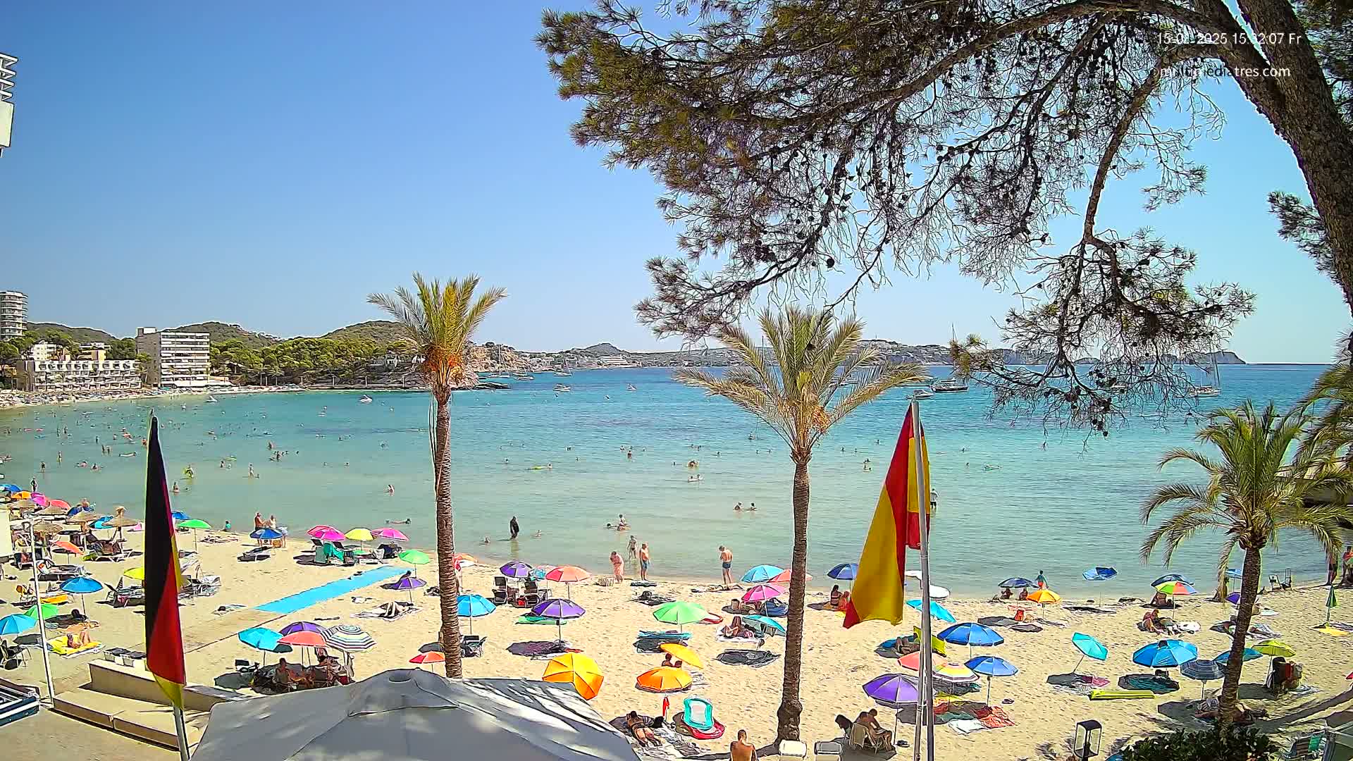 A sunny beach scene shows a sandy shore with people swimming and sunbathing, palm trees, and boats on a calm, clear blue bay, under a mostly clear sky.