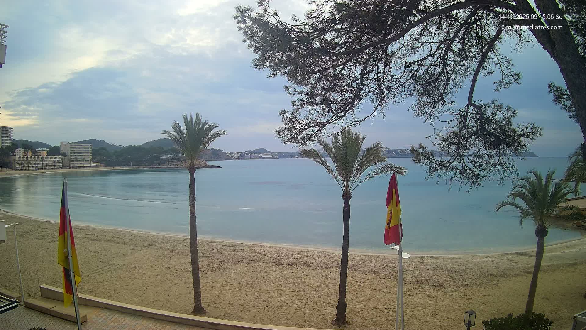 A sunny day with scattered clouds lights up a sandy beach with palm trees and a German and Spanish flag, overlooking a bay with gentle waves and distant coastal buildings and hills.