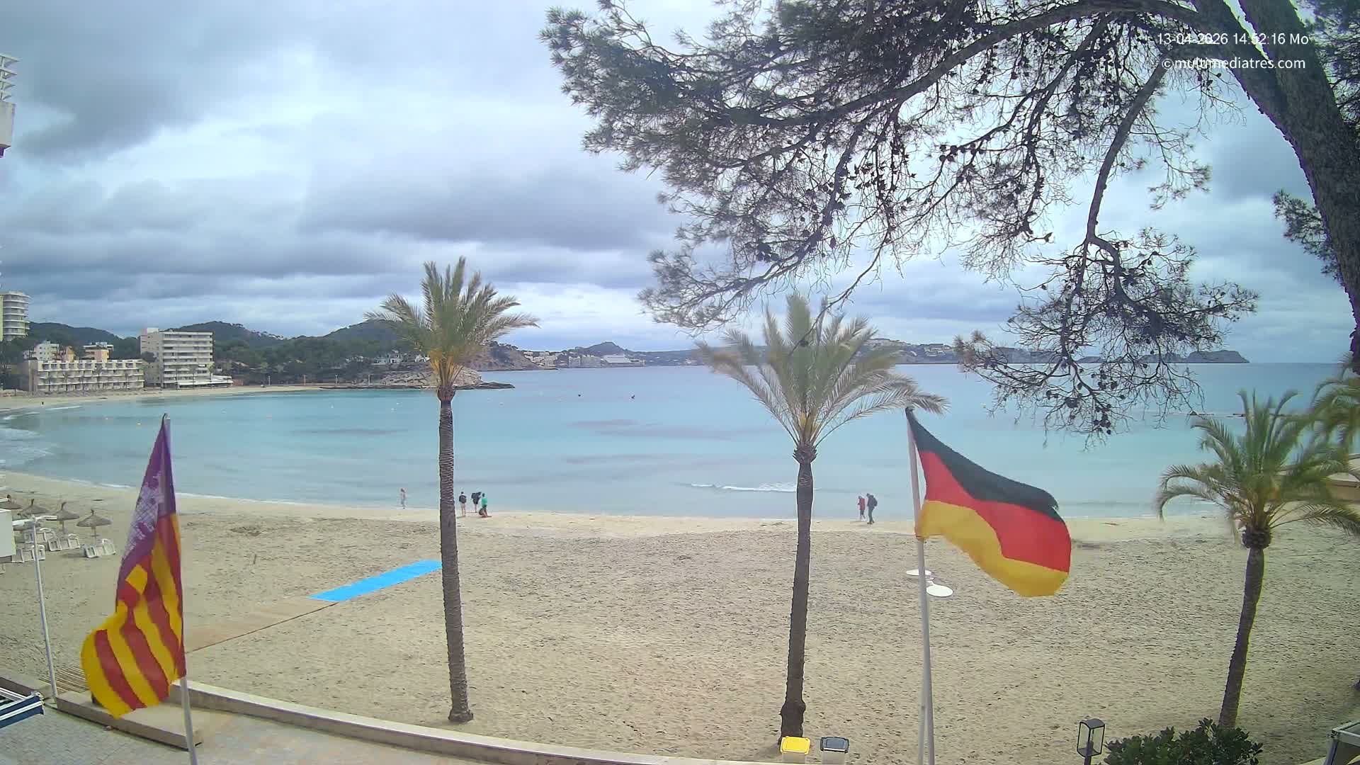 A sunny day with scattered clouds lights up a sandy beach with palm trees and a German and Spanish flag, overlooking a bay with gentle waves and distant coastal buildings and hills.