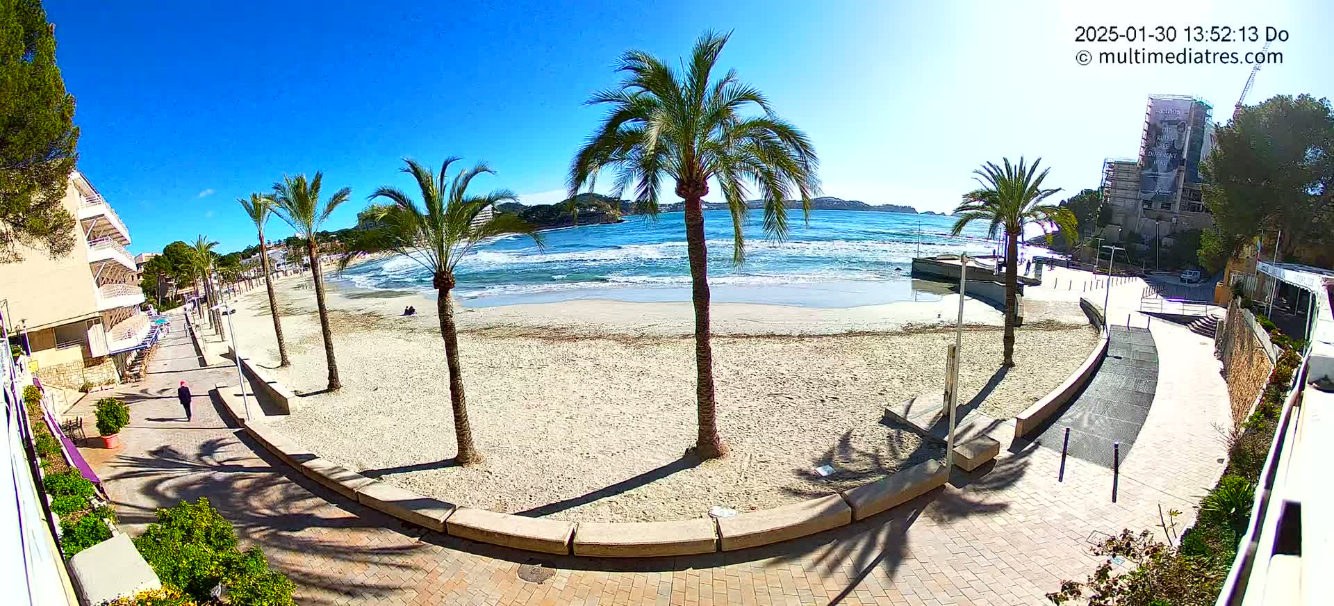 A sandy beach with palm trees and a paved walkway next to a building, under sunny skies with some small waves in the ocean.