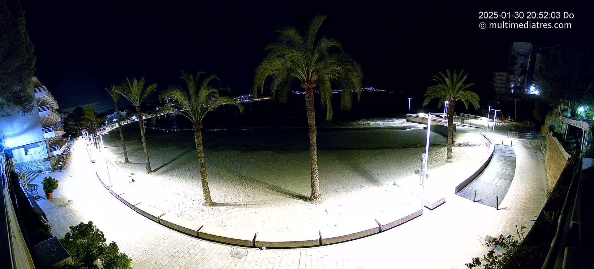 A nighttime, brightly lit beachside promenade curves around a sandy beach, featuring several palm trees and buildings in the background under a clear, dark sky.