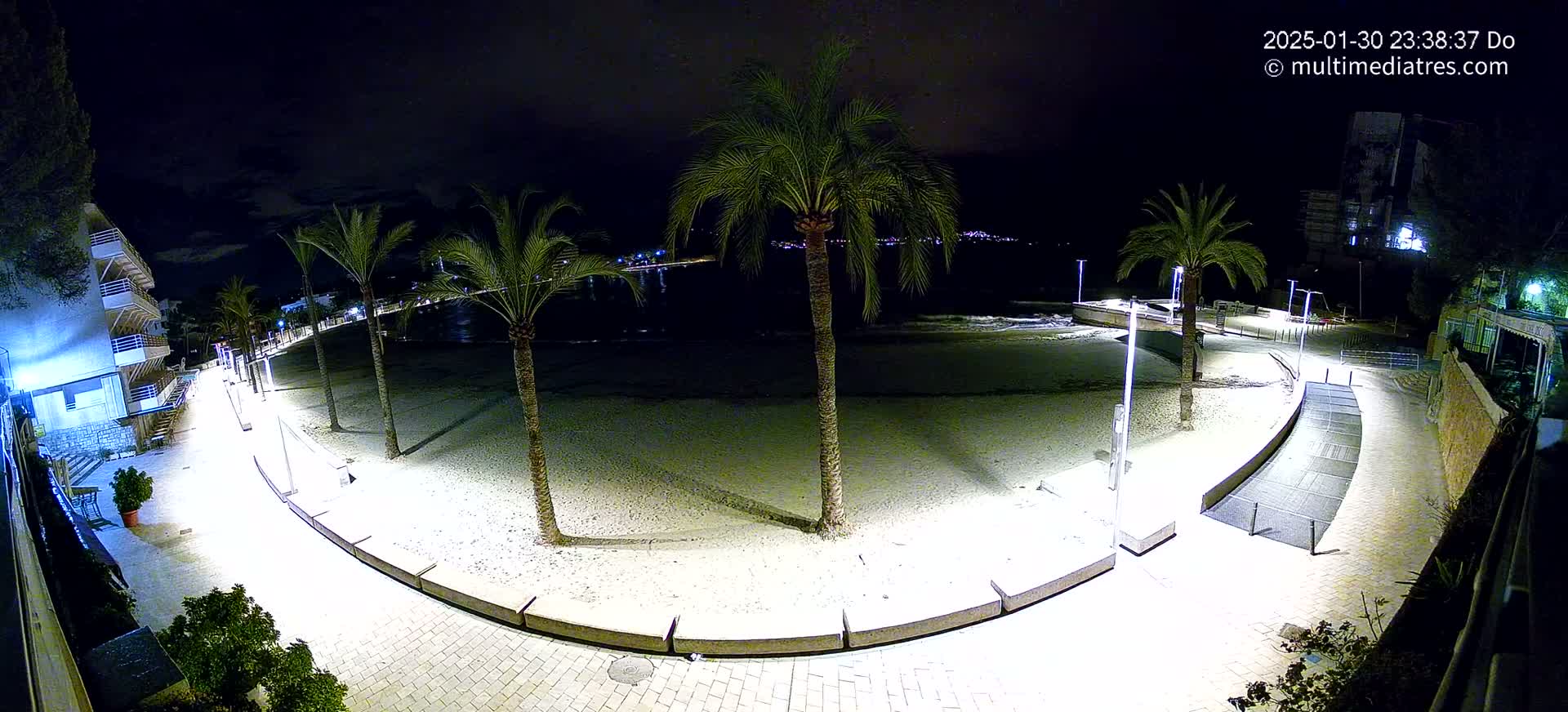 A nighttime view of a sandy beach, lined with palm trees and illuminated walkways, overlooking a calm sea.