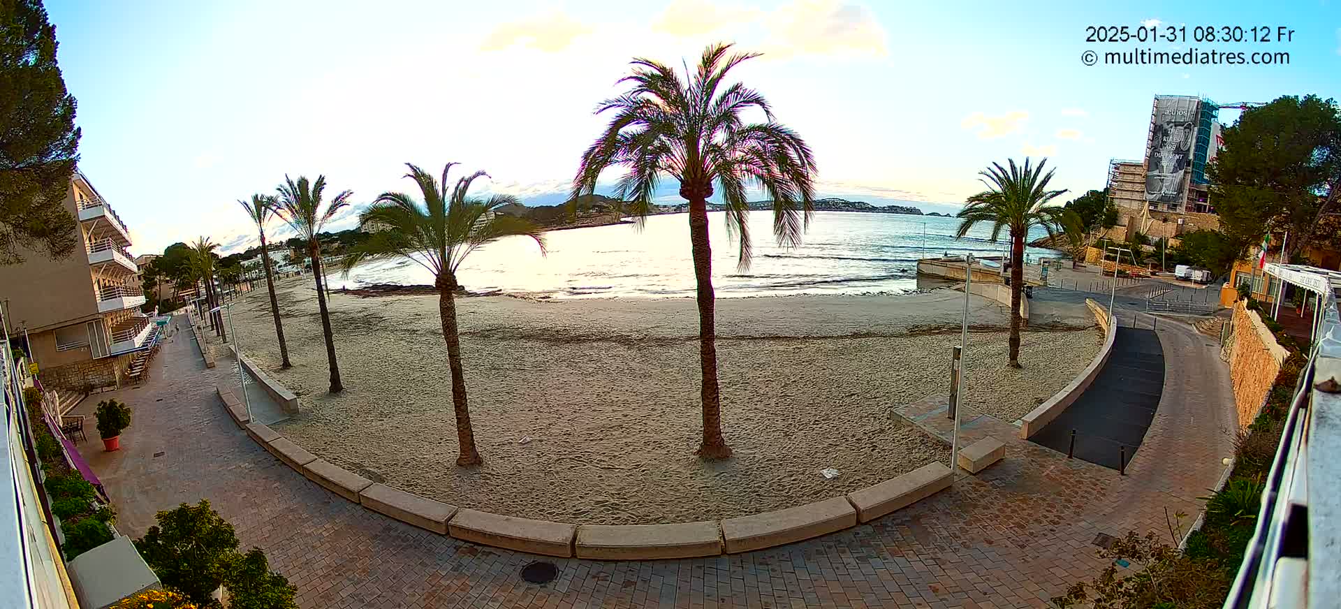 A sandy beach with palm trees is visible next to a paved walkway leading to buildings, under a partly cloudy sky.