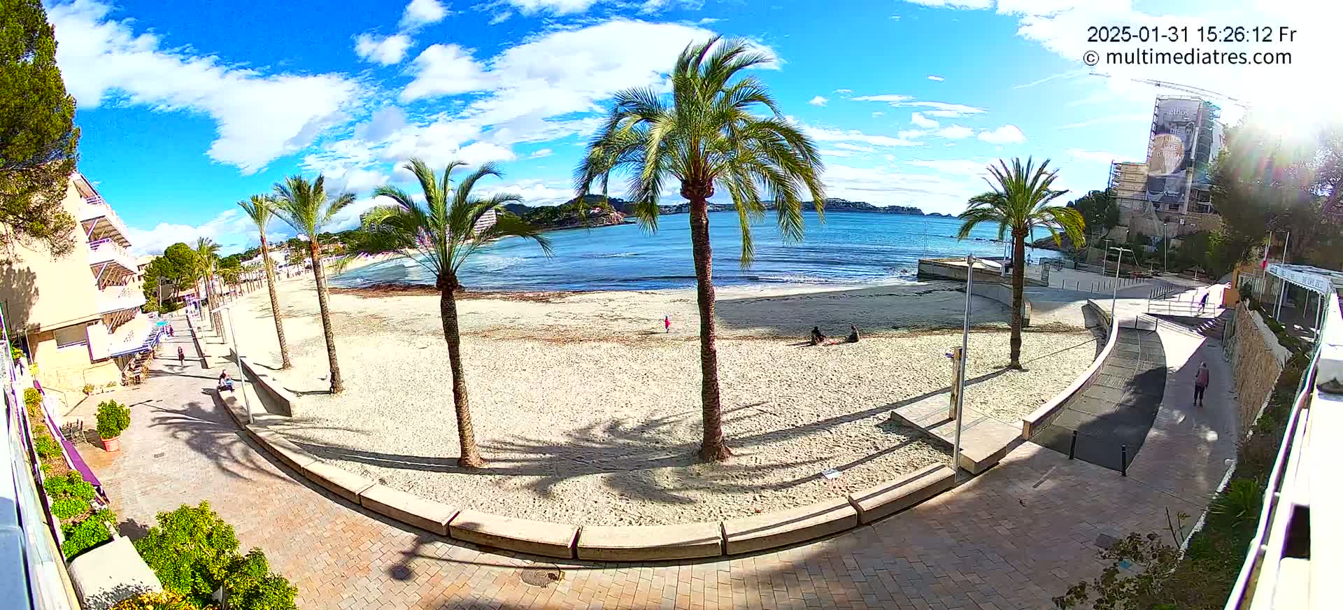 A mostly sunny day at a sandy beach, with palm trees lining a paved walkway next to calm blue water and a few people sparsely scattered on the sand.