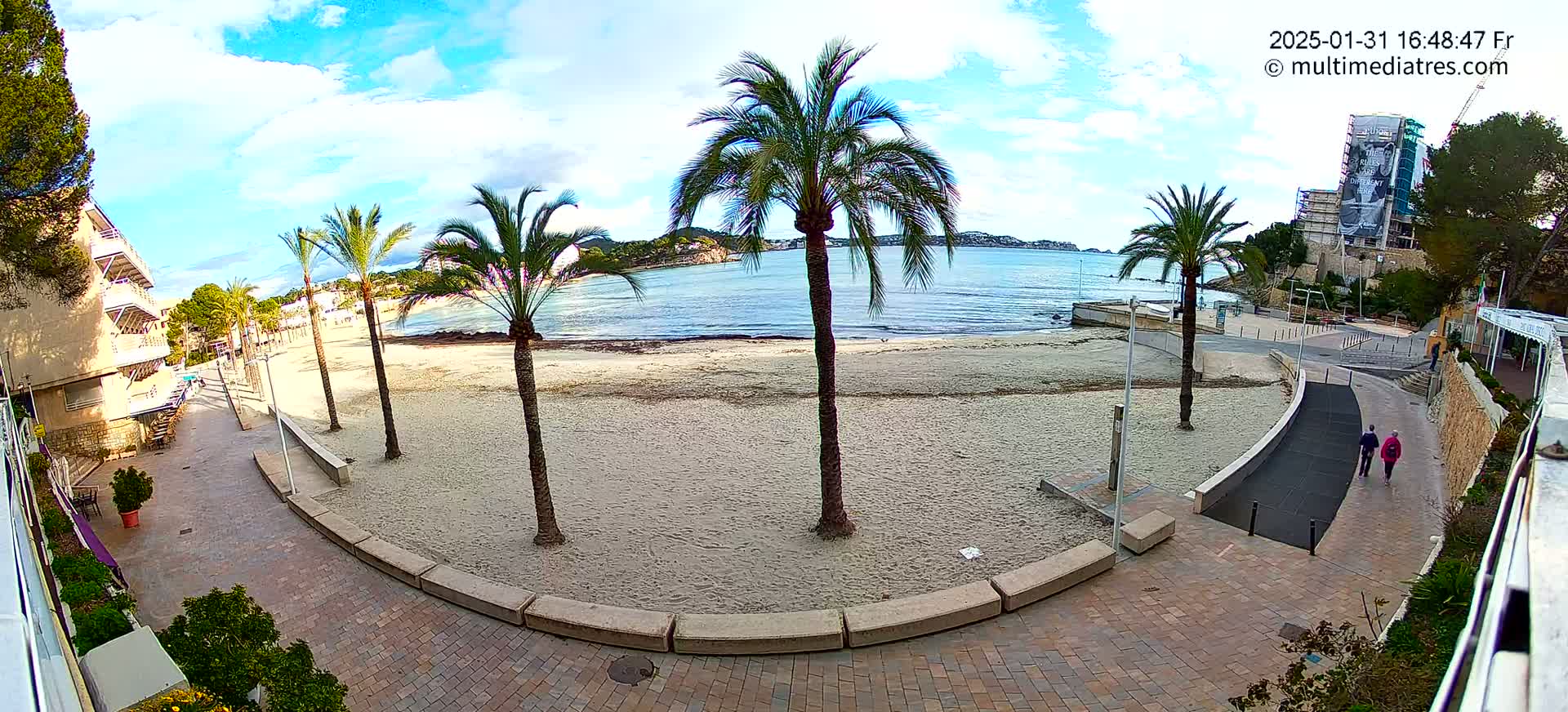 A mostly sunny day at a sandy beach with palm trees, a paved walkway, and a few people walking.
