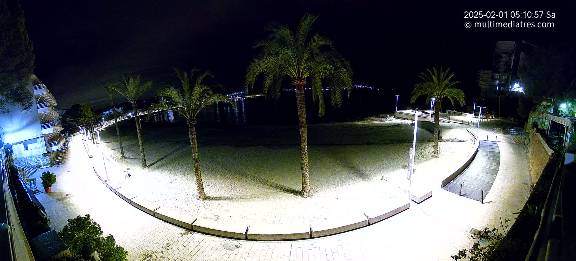 A nighttime, brightly lit beach scene shows a curving paved walkway lined with palm trees, leading to a sandy beach and calm water with distant lights.