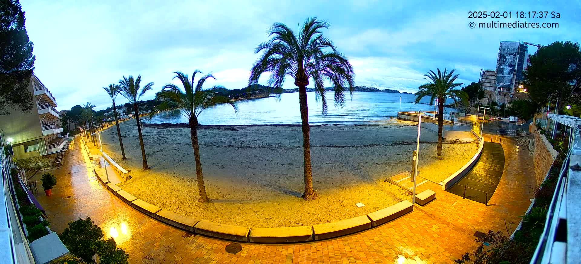 A wet, illuminated beachfront promenade curves around a sandy beach at dusk, lined with palm trees and buildings.