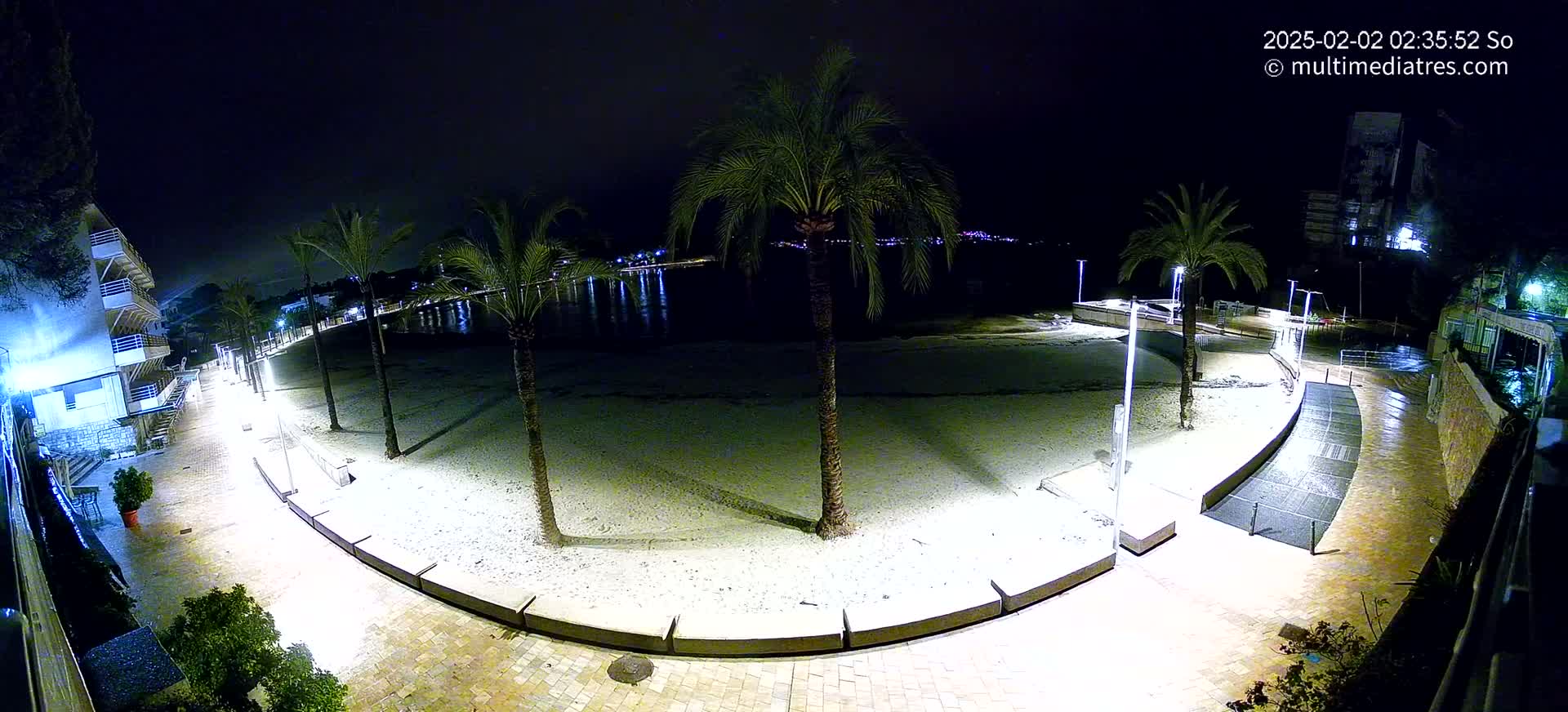 A nighttime, illuminated view of a sandy beach bordered by a paved walkway, palm trees, and buildings, with calm water visible in the background.
