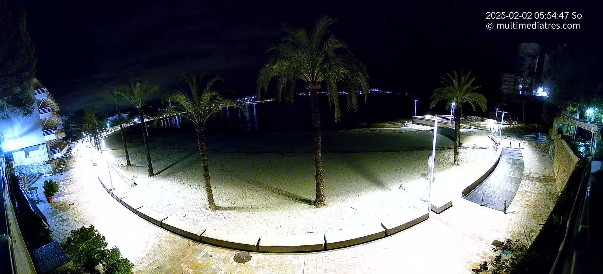 A nighttime view of a crescent-shaped beach, illuminated by lights, lined with palm trees and bordered by a paved walkway.