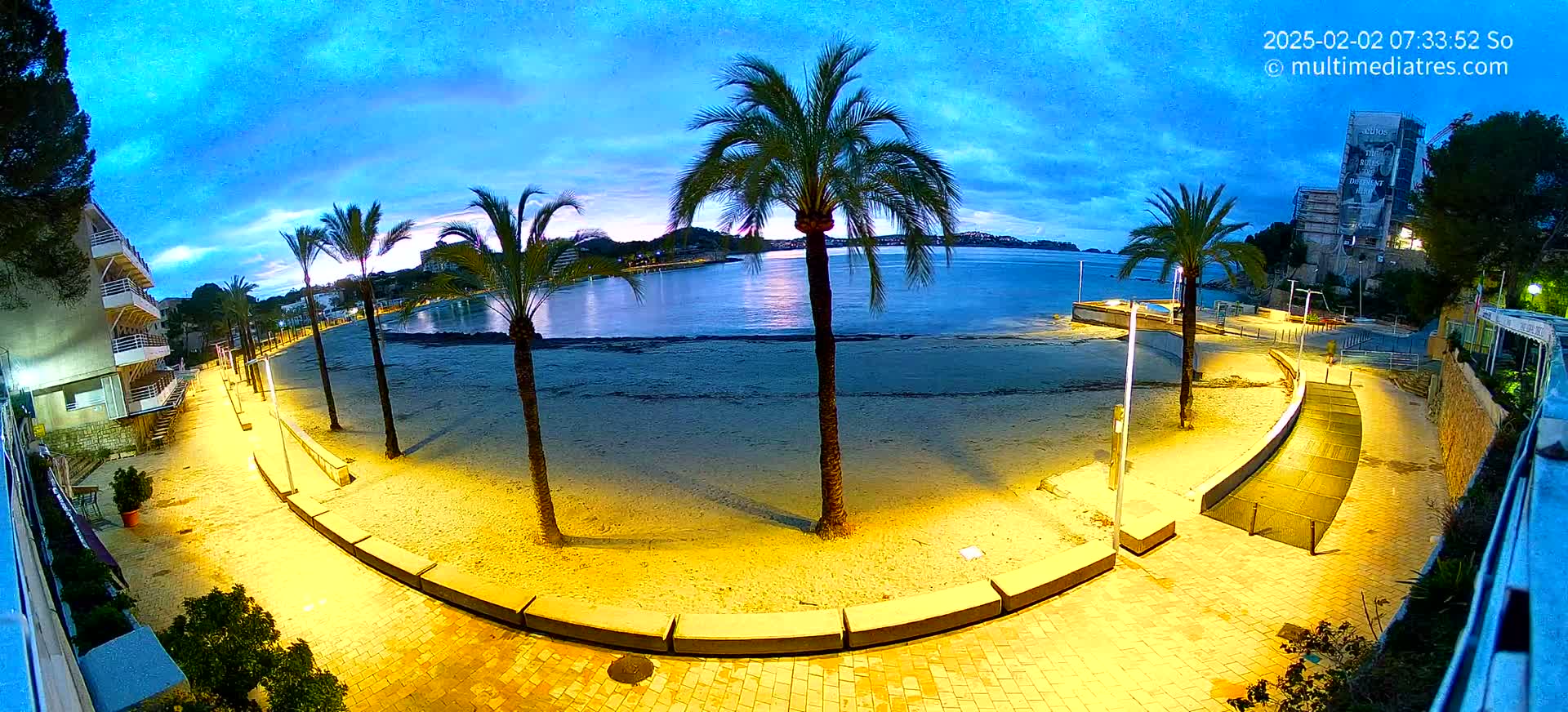A sandy beach at night is illuminated by yellow lights along a paved walkway, bordered by palm trees and buildings under a twilight sky.