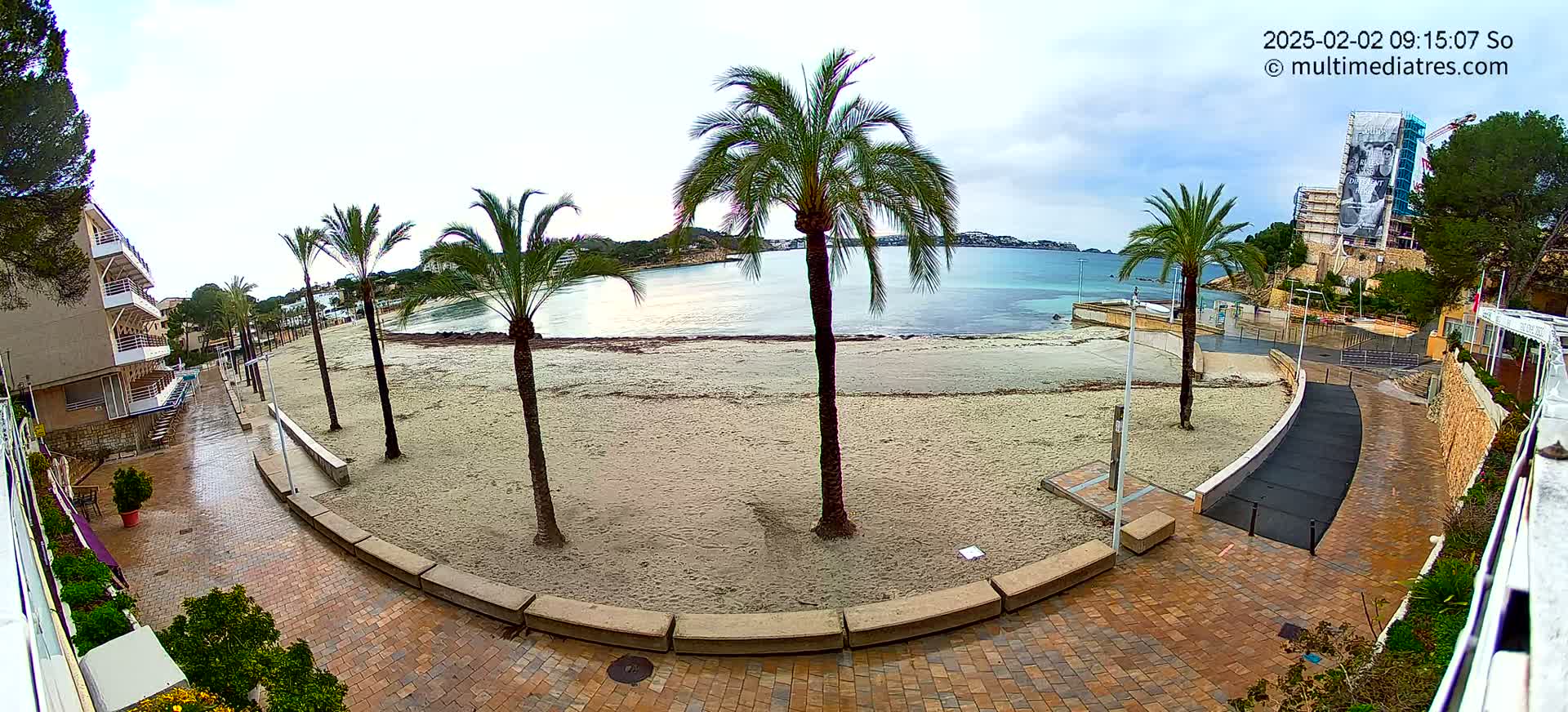 A wet, sandy beach with several palm trees is visible from a brick paved walkway overlooking a calm body of water under an overcast sky.