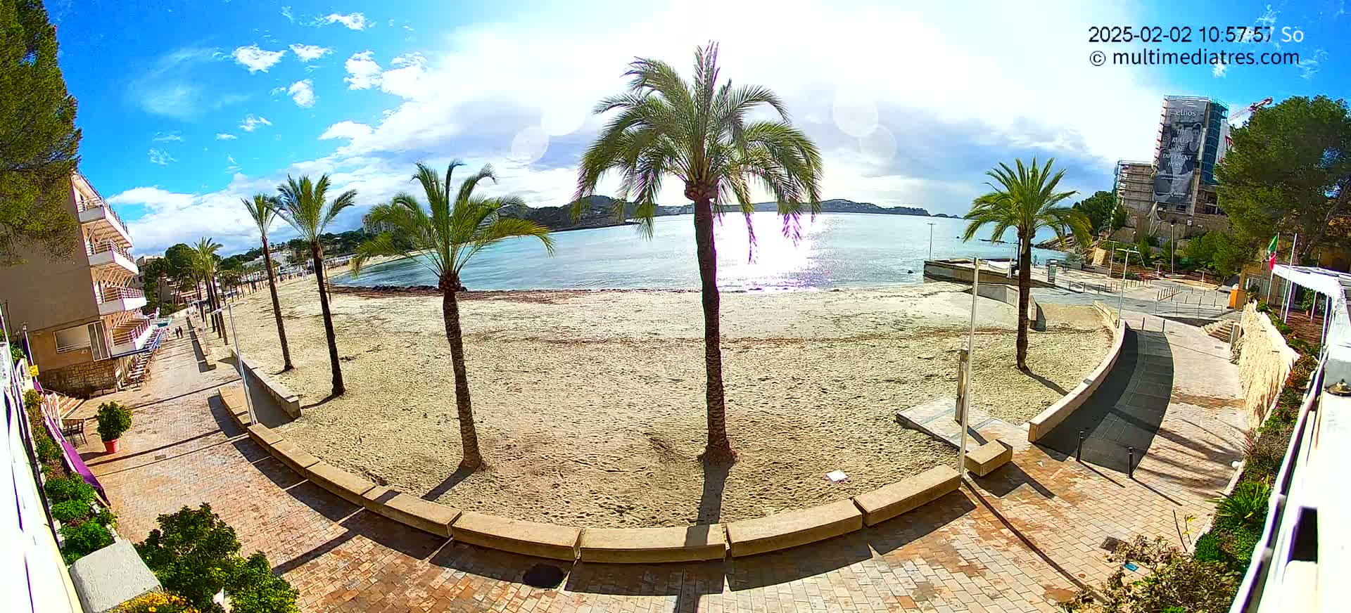 A mostly sunny day shows a sandy beach with palm trees, a paved walkway, and buildings nearby.