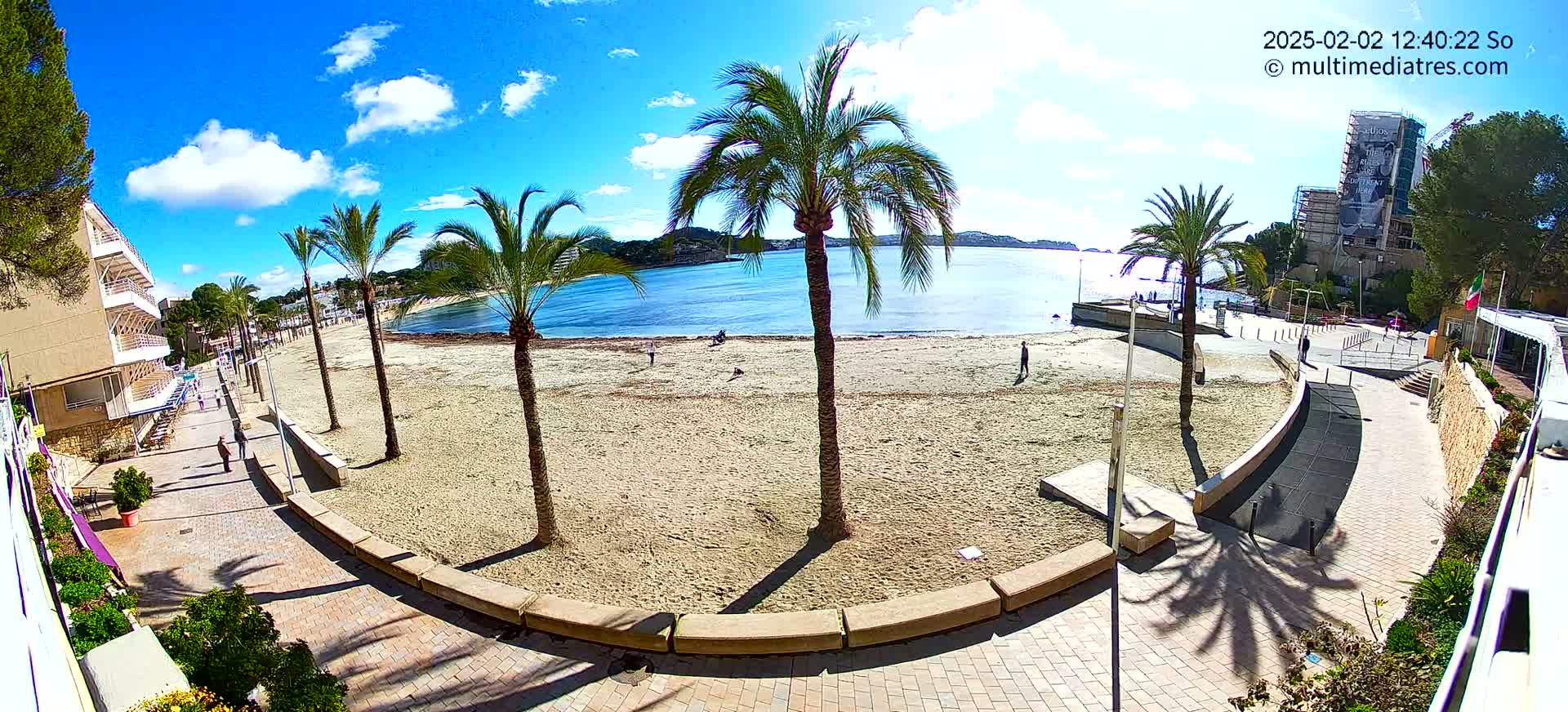 A sandy beach with palm trees and a paved walkway overlooks a calm bay under a sunny sky with some clouds.