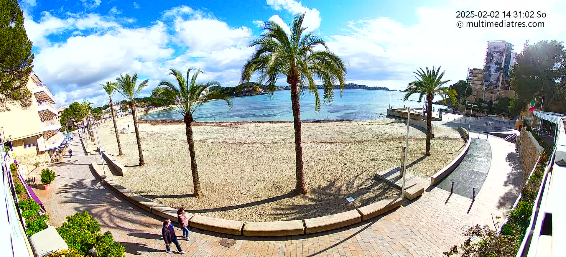 A sandy beach with palm trees and a paved walkway borders a calm, blue body of water under a partly cloudy sky.