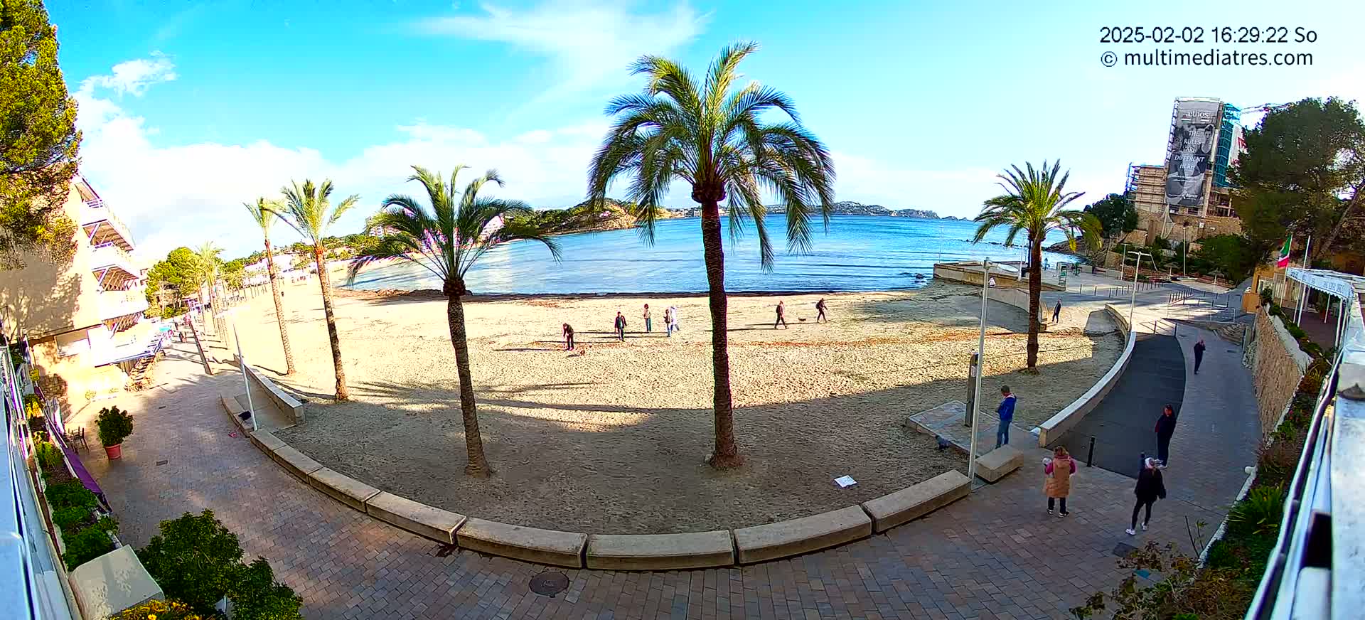 A sandy beach with palm trees and a few people walking along the shore on a sunny day.