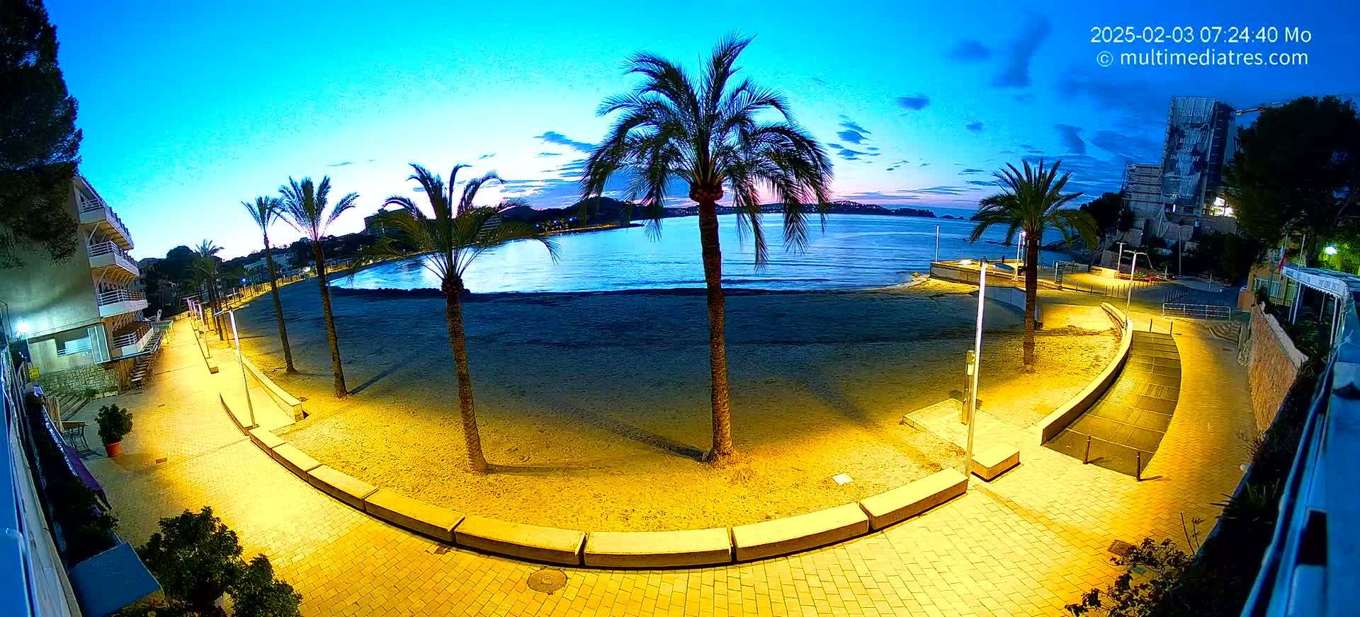 A sandy beach at twilight is illuminated by yellow lights, lined with palm trees and a paved walkway leading to buildings.