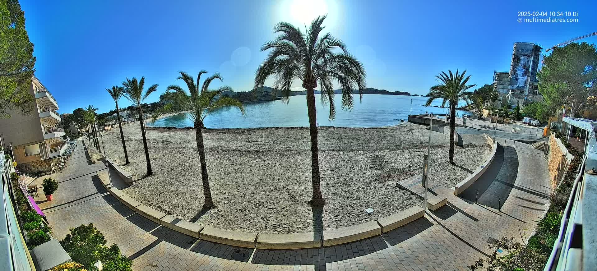 A sandy beach with palm trees lines a paved walkway next to calm, blue water under a sunny sky.