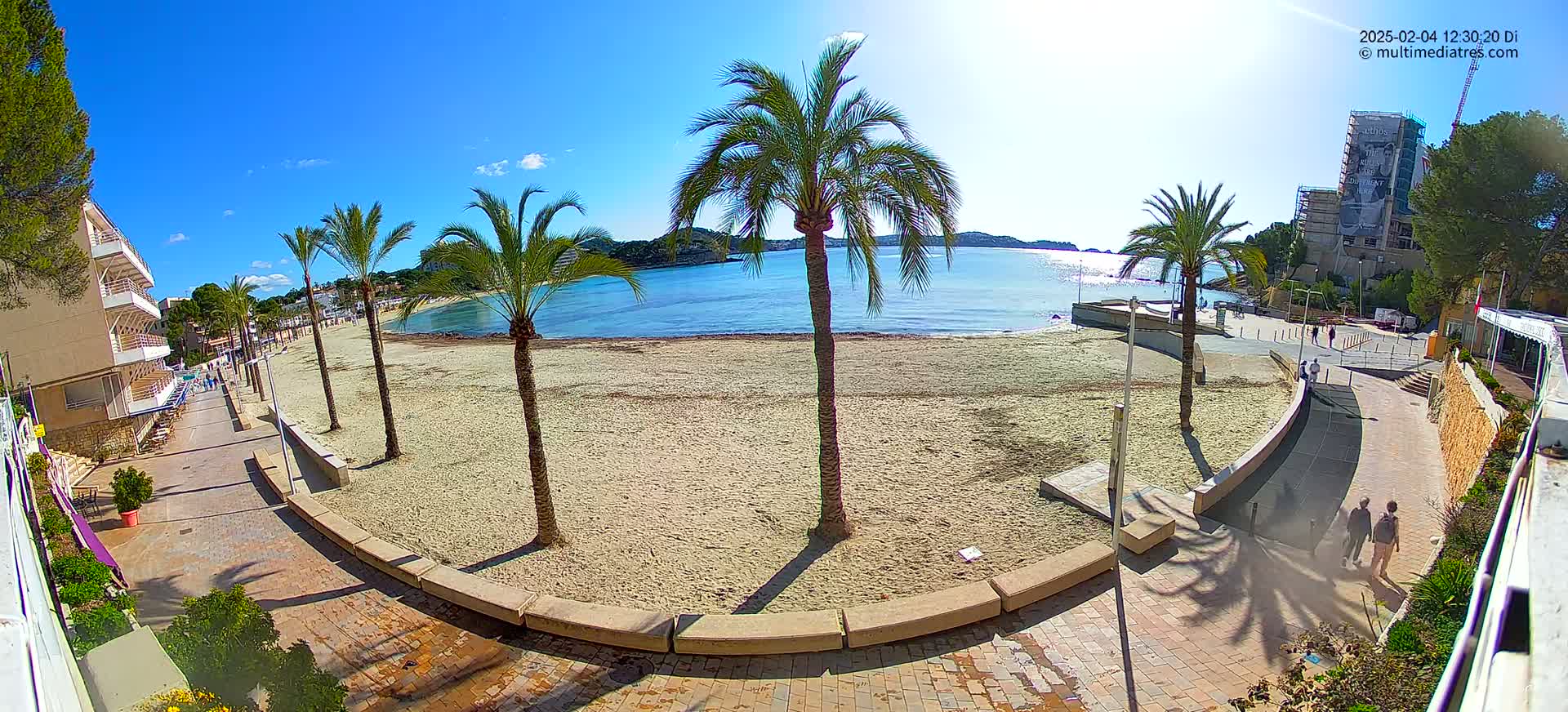 A sandy beach with palm trees and a paved walkway overlooks a calm bay on a sunny day.