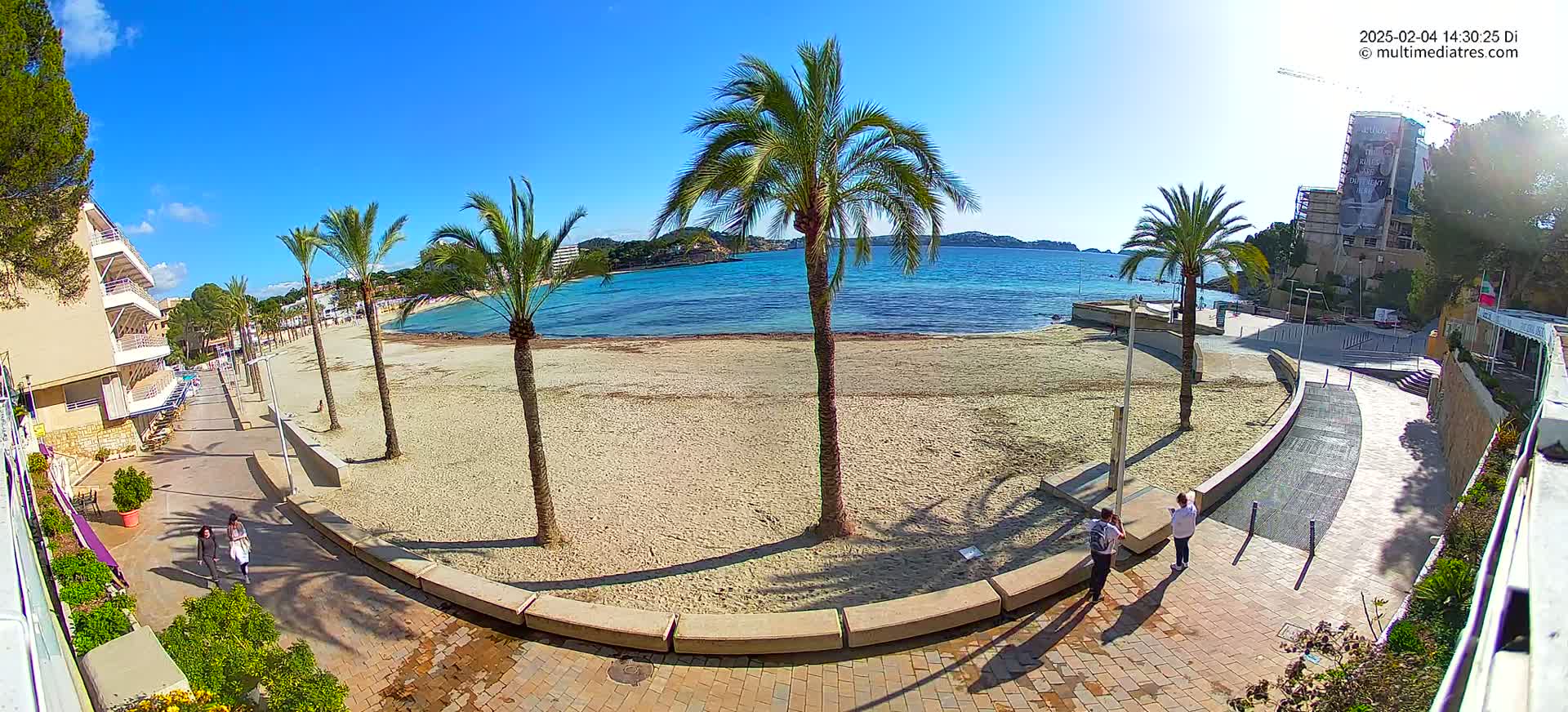 A sandy beach with palm trees and buildings is seen on a sunny day, with people walking along a paved walkway next to the beach.
