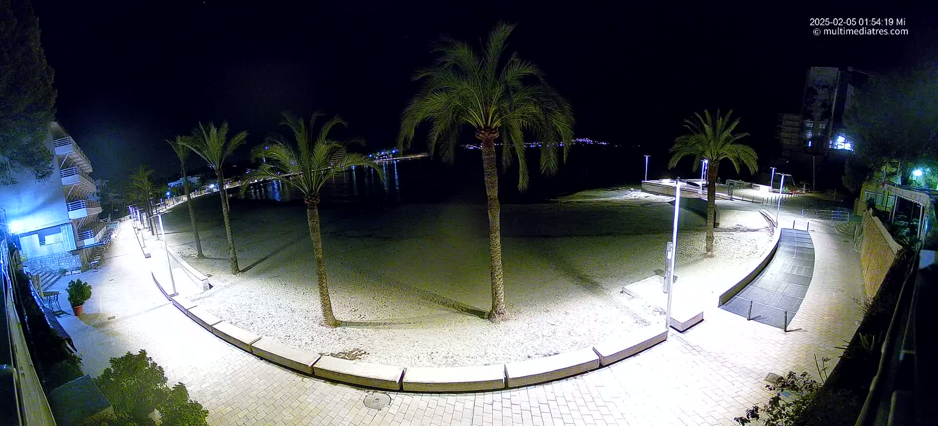 A paved walkway curves around a sandy beach at night, illuminated by artificial lights, with palm trees lining the beachfront and buildings visible in the background.