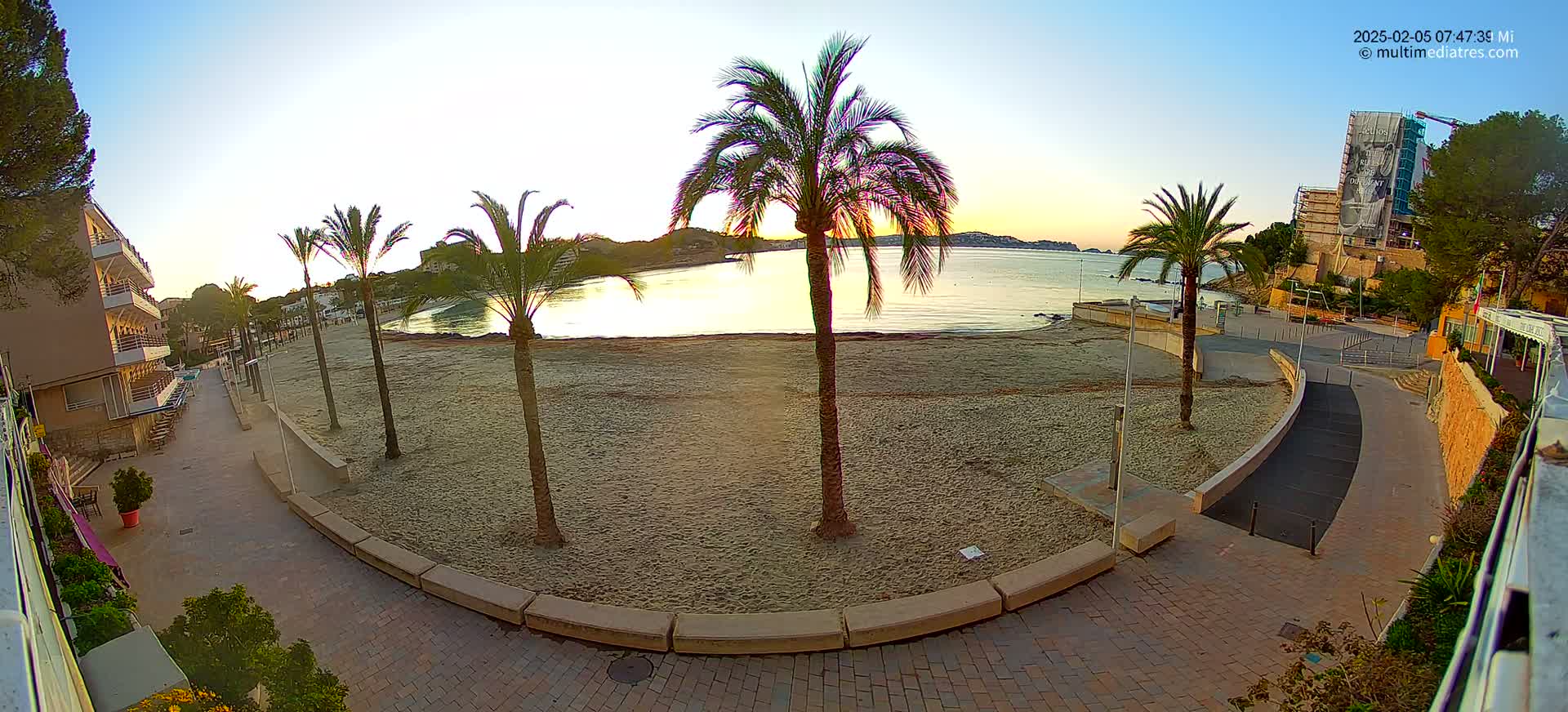 A sandy beach at sunrise, lined with palm trees and a paved walkway, overlooks calm water and buildings.