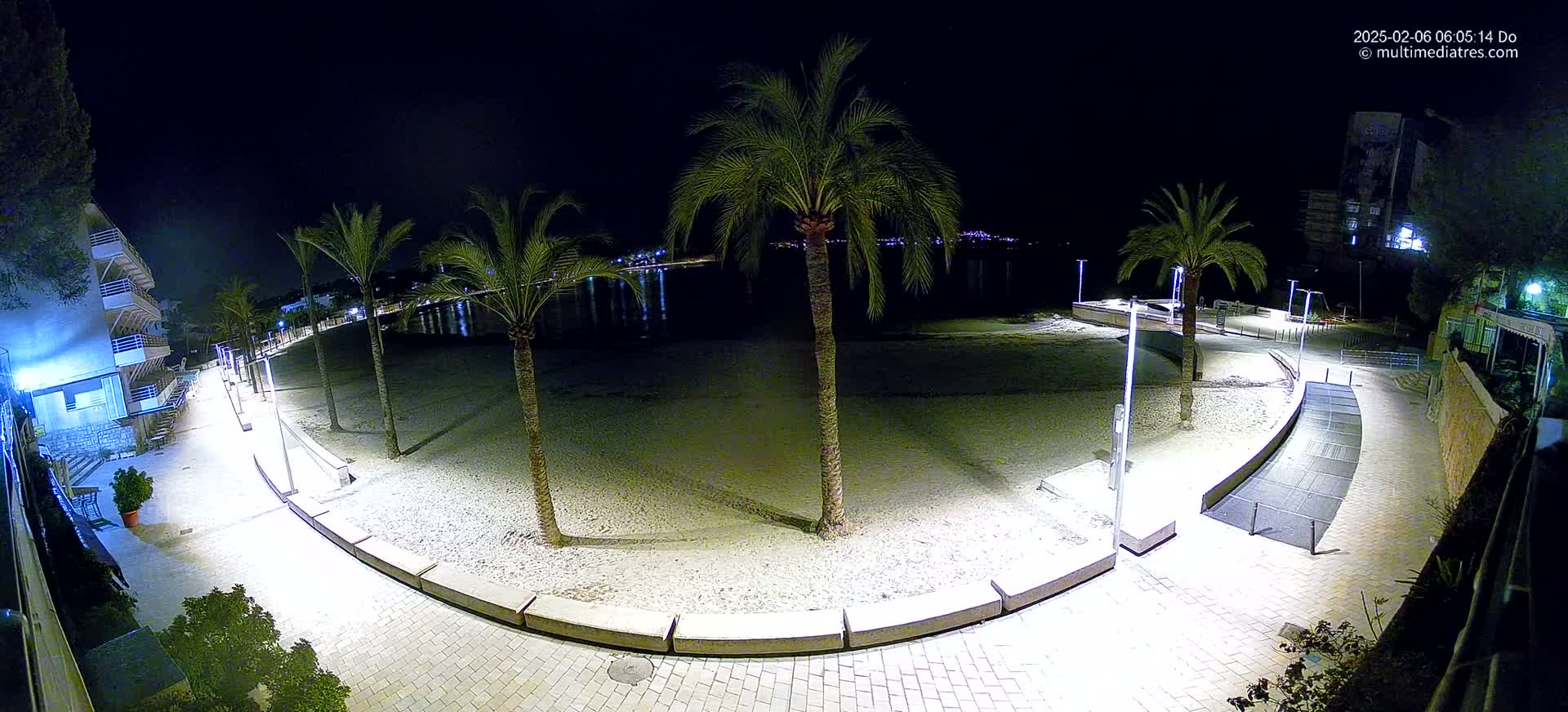 A nighttime view of a sandy beach with palm trees, illuminated walkways, and buildings in the background under a clear, dark sky.