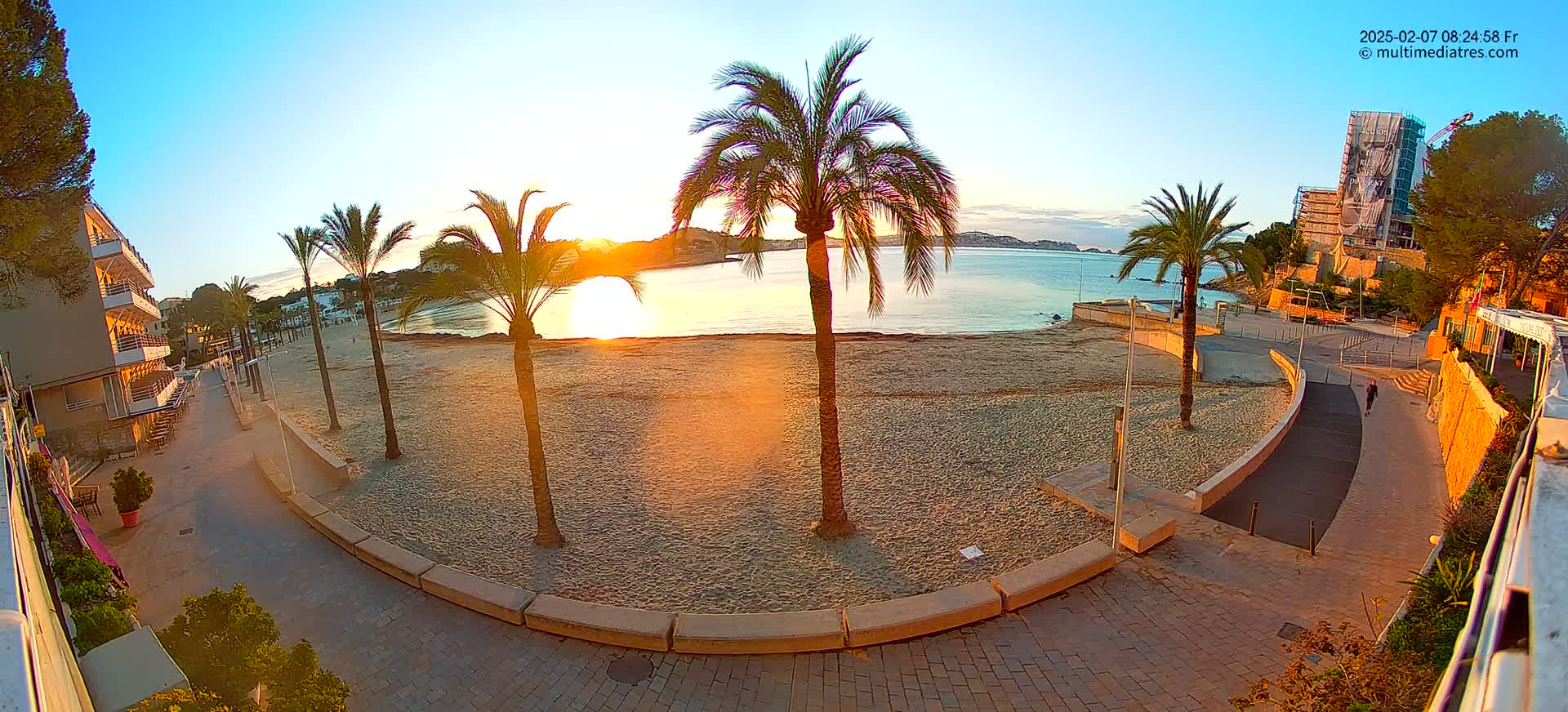 A sandy beach at sunrise, lined with palm trees and a paved walkway, overlooks calm water and distant buildings.