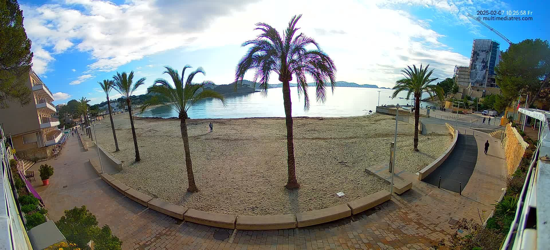 A mostly sunny day reveals a sandy beach with palm trees lining a paved walkway leading to buildings and a calm body of water.