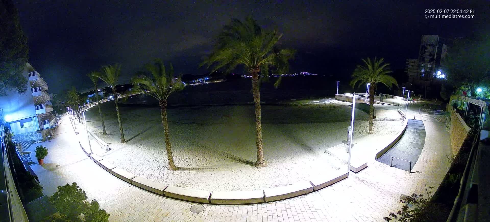 A nighttime, high-angle view shows a sandy beach bordered by a paved walkway lined with palm trees and illuminated by streetlights, overlooking a calm body of water and a distant town.
