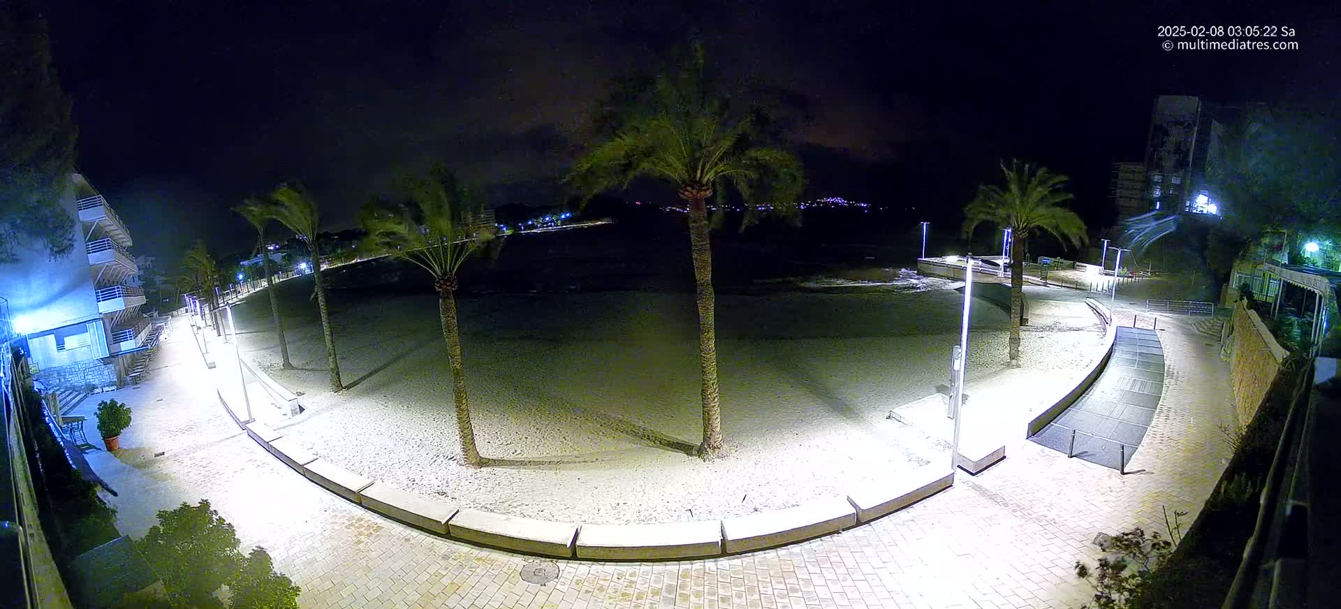 A nighttime view of a sandy beach with palm trees, lit walkways, and buildings in the background, under a clear, dark sky.