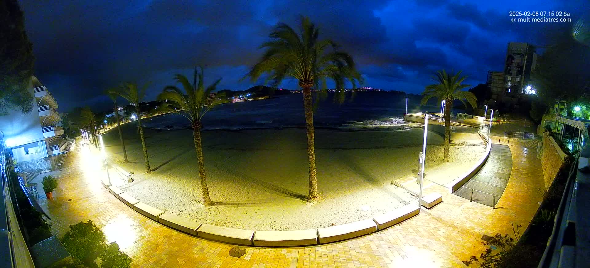 A nighttime view of a sandy beach with palm trees and a paved walkway, illuminated by lights, under a partly cloudy sky.