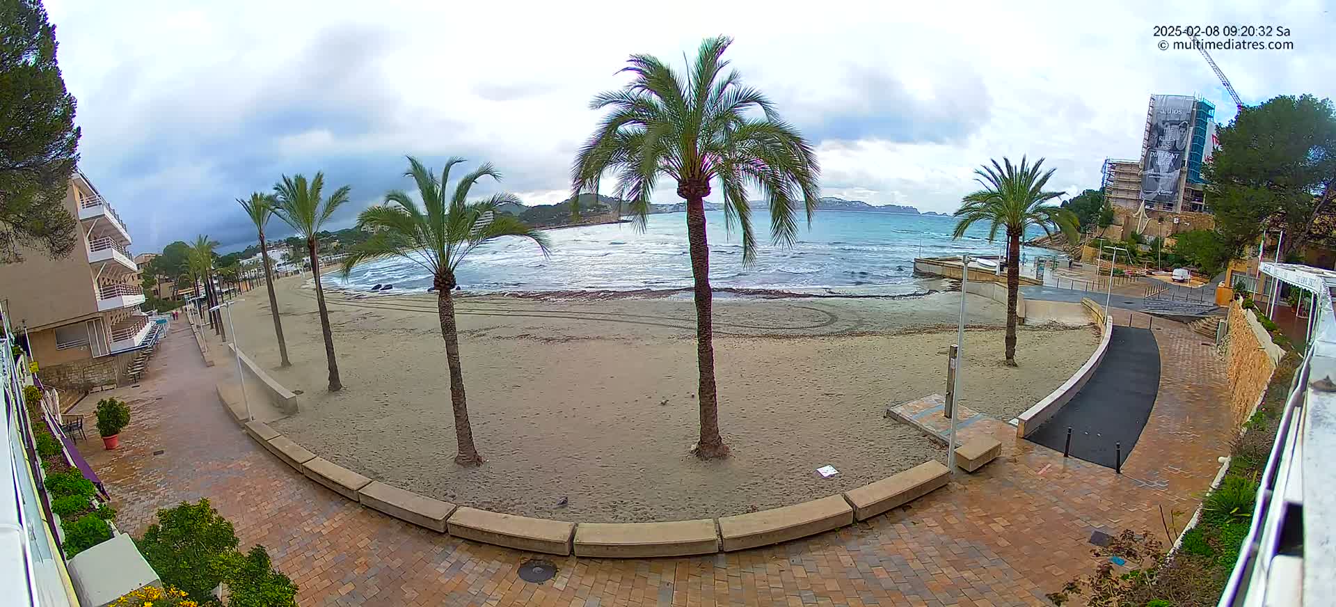 A sandy beach with several palm trees is visible, overlooking a somewhat choppy sea under a cloudy sky.