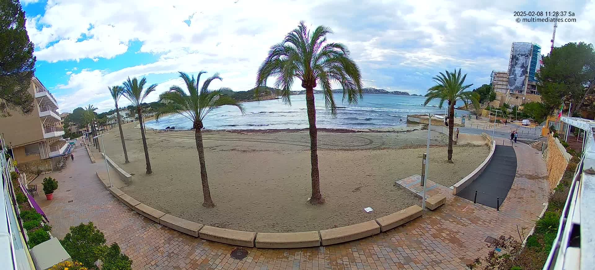 A sandy beach with palm trees and a paved walkway is seen under a partly cloudy sky, with a calm sea in the background.