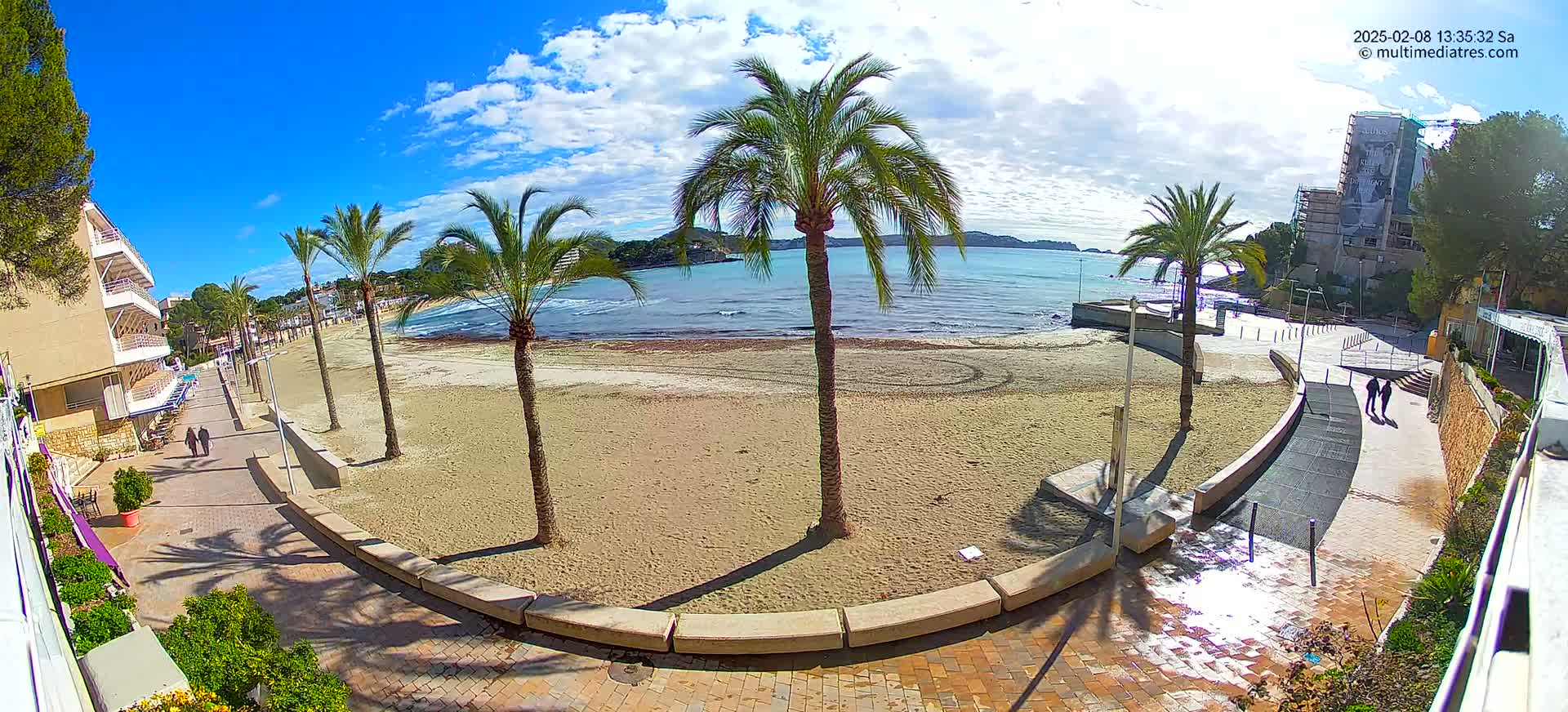 A sandy beach with palm trees and a paved walkway borders a calm, blue ocean under a mostly sunny sky.