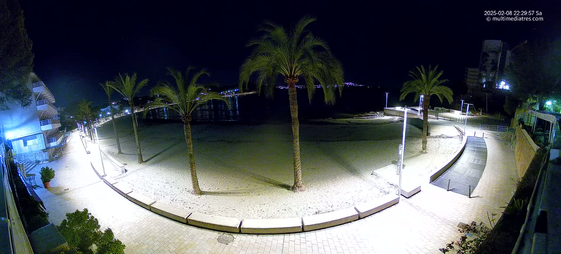 A nighttime view of a sandy beach bordered by a paved walkway lined with palm trees and lights, overlooking a calm body of water and a distant town.