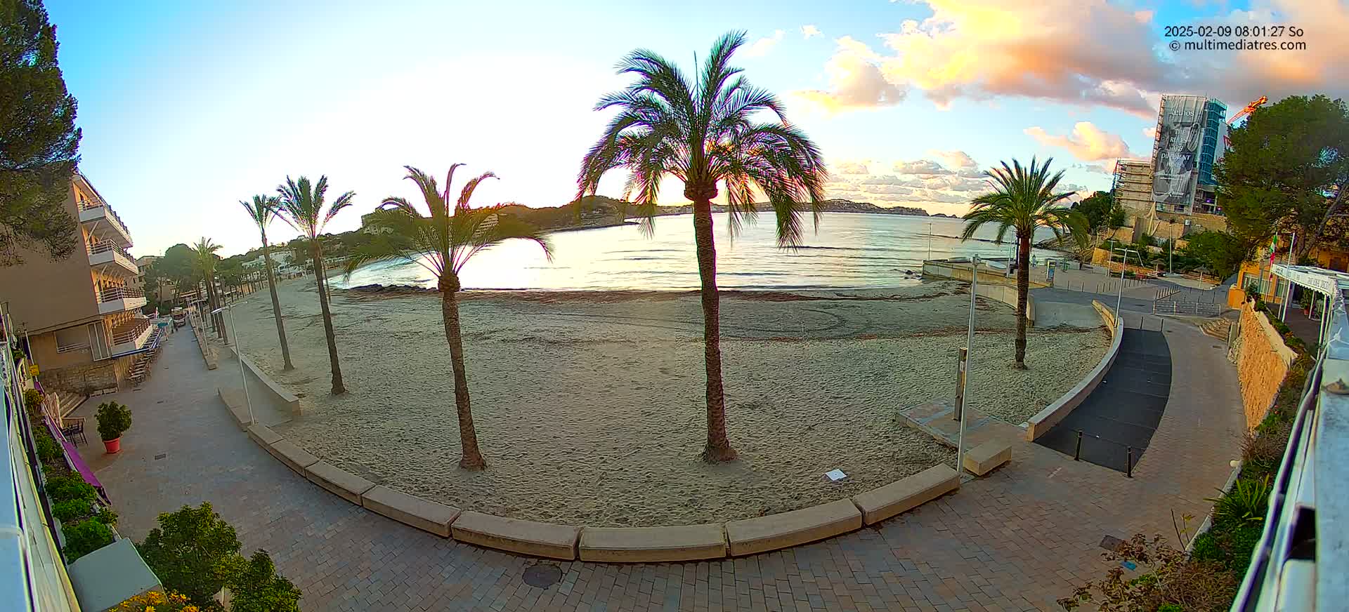 A sandy beach at sunrise is bordered by a paved walkway, palm trees, and buildings.