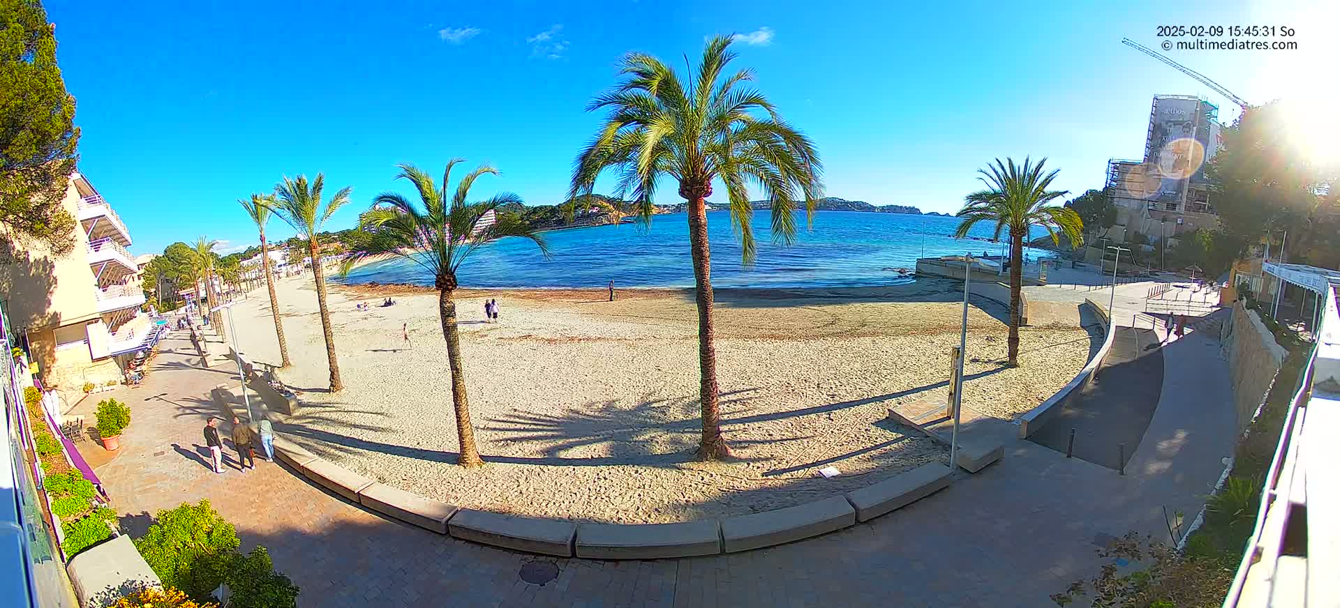 A sandy beach with palm trees and buildings is visible, under a clear blue sky and bright sunshine.