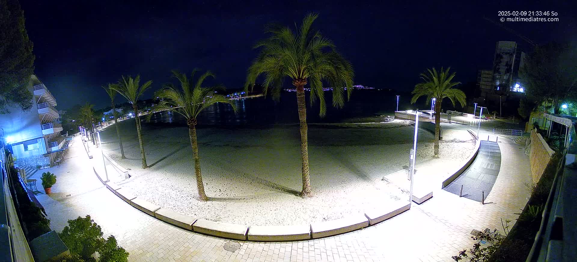 An illuminated sandy beach at night, lined with palm trees and a paved walkway, overlooks a calm body of water with city lights visible in the distance.