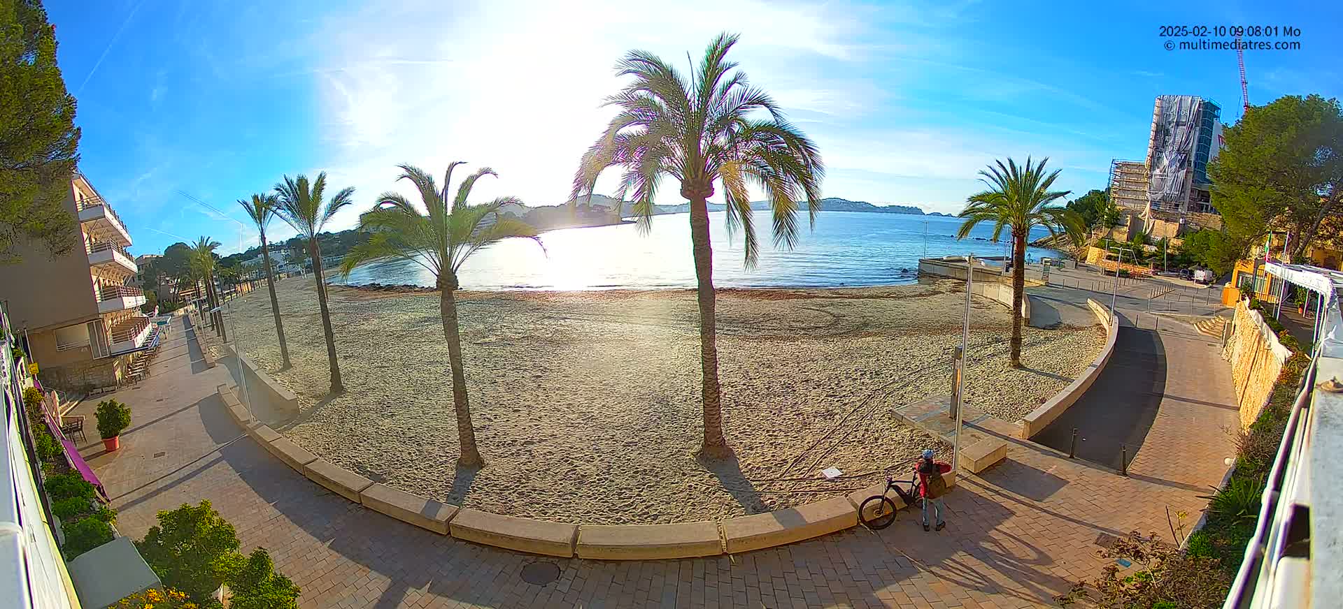 A sandy beach with palm trees and a promenade, under a clear sunny sky, is viewed from above, showing a person with a bicycle near a building under construction in the distance.