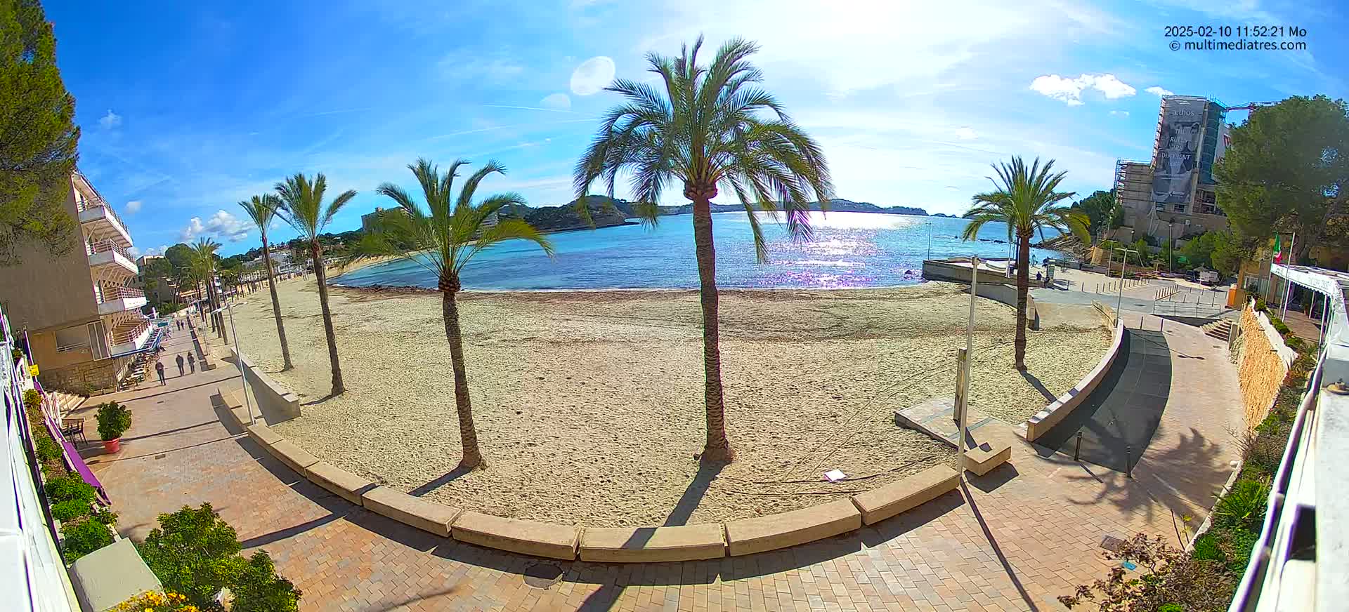 A sandy beach with palm trees lines a paved walkway next to a calm, sunny sea, under a mostly clear blue sky.