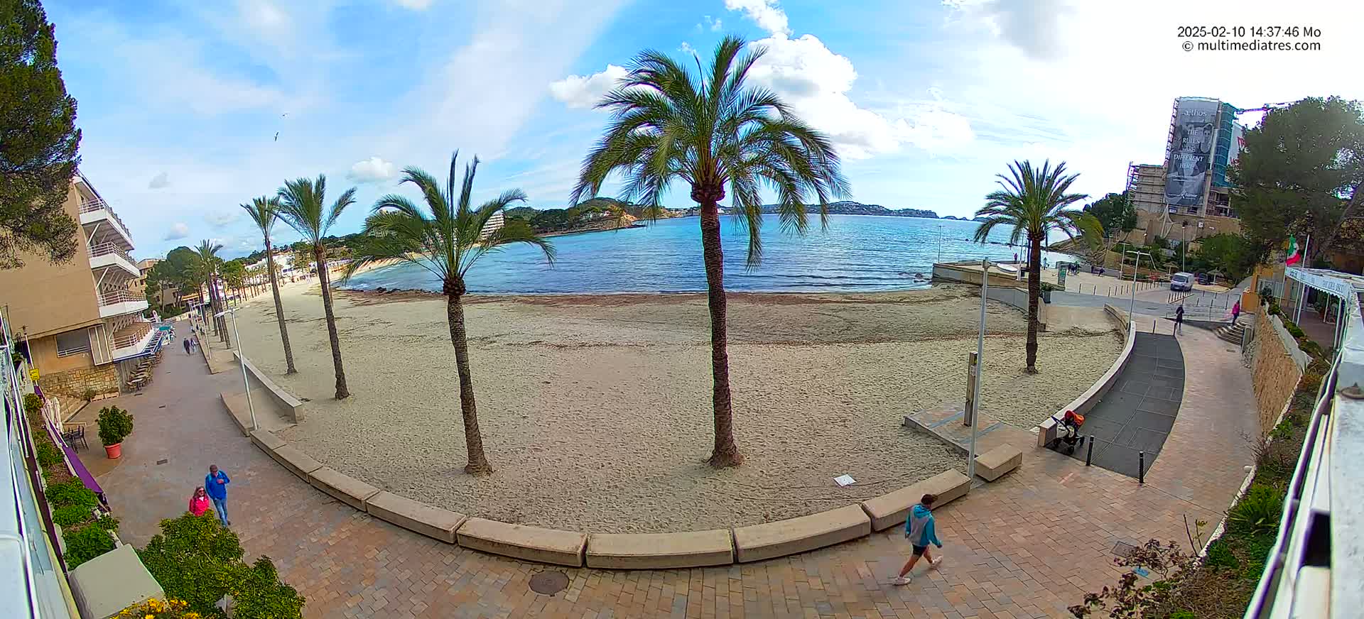 A sandy beach with palm trees is visible from a paved walkway overlooking a calm, blue body of water under a partly cloudy sky.