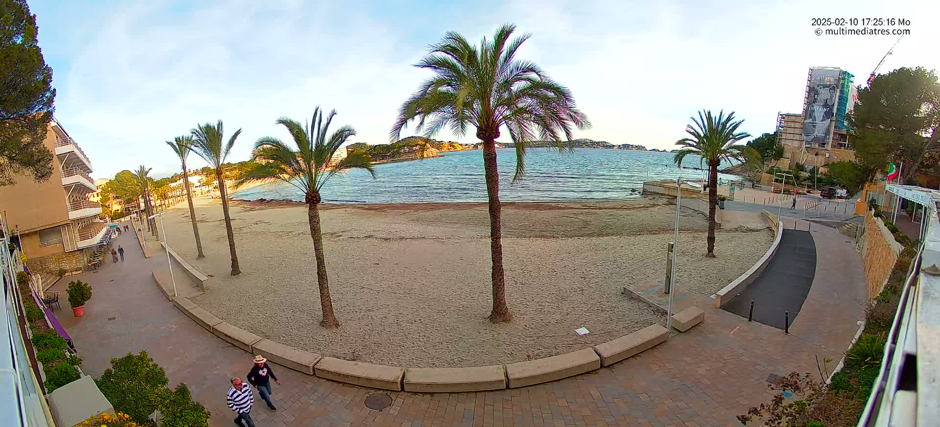 A mostly sunny beach scene shows a sandy beach, palm trees, buildings, and a paved walkway with a few people walking.