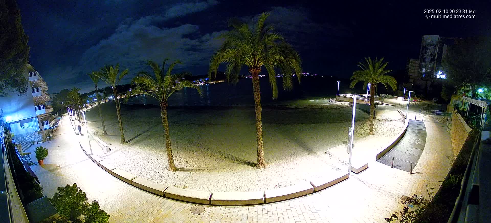A nighttime view of a sandy beach with palm trees and a paved walkway, illuminated by artificial lights under a partly cloudy sky.