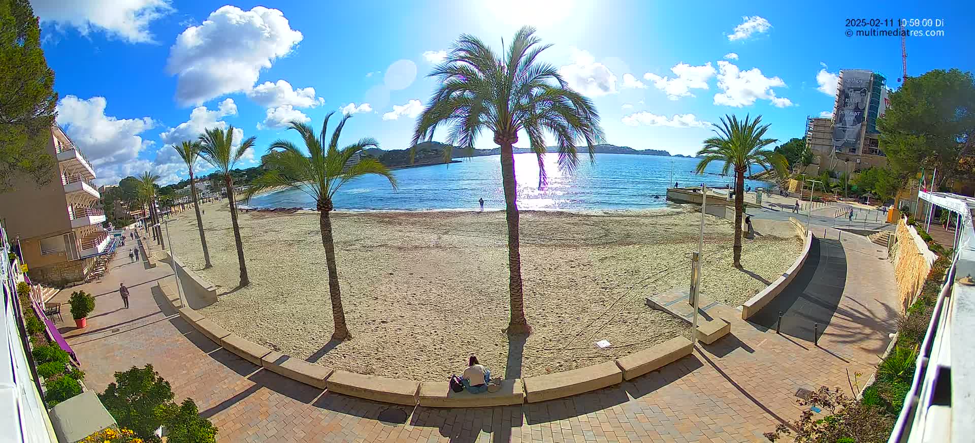 A sandy beach with palm trees lines a paved walkway next to calm, sunlit water under a mostly sunny sky.